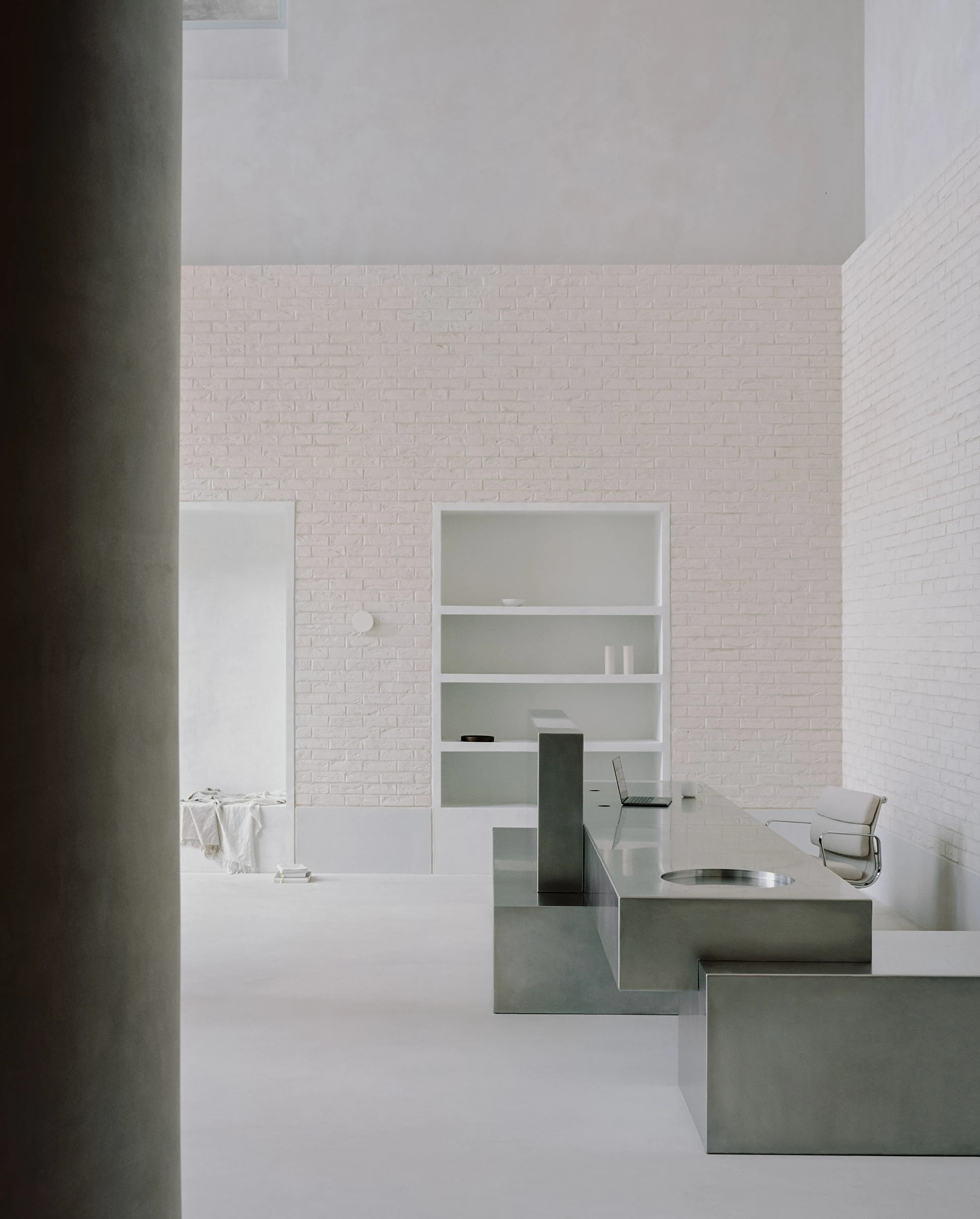 Cherry Orchard by Actual Office, photographed by Nick Dearden — Wider view of the entrance hall from behind the plastered column: stainless concierge desk in the foreground with an integrated drop-point bowl, white brick wall and recessed open shelf, a…