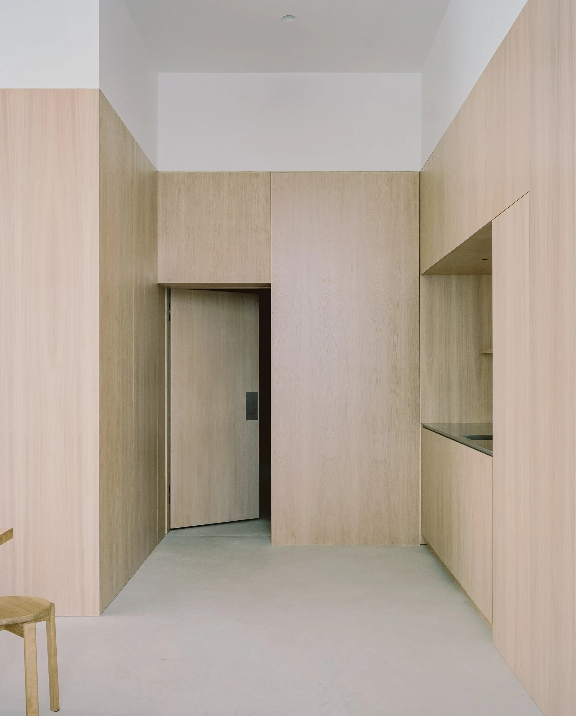 Cherry Orchard by Actual Office, photographed by Nick Dearden — Communal room looking back toward an oak-clad service wall: stainless-steel kitchen worktop on the left, a square internal aperture glazed in opaque green at the centre, ash communal table…