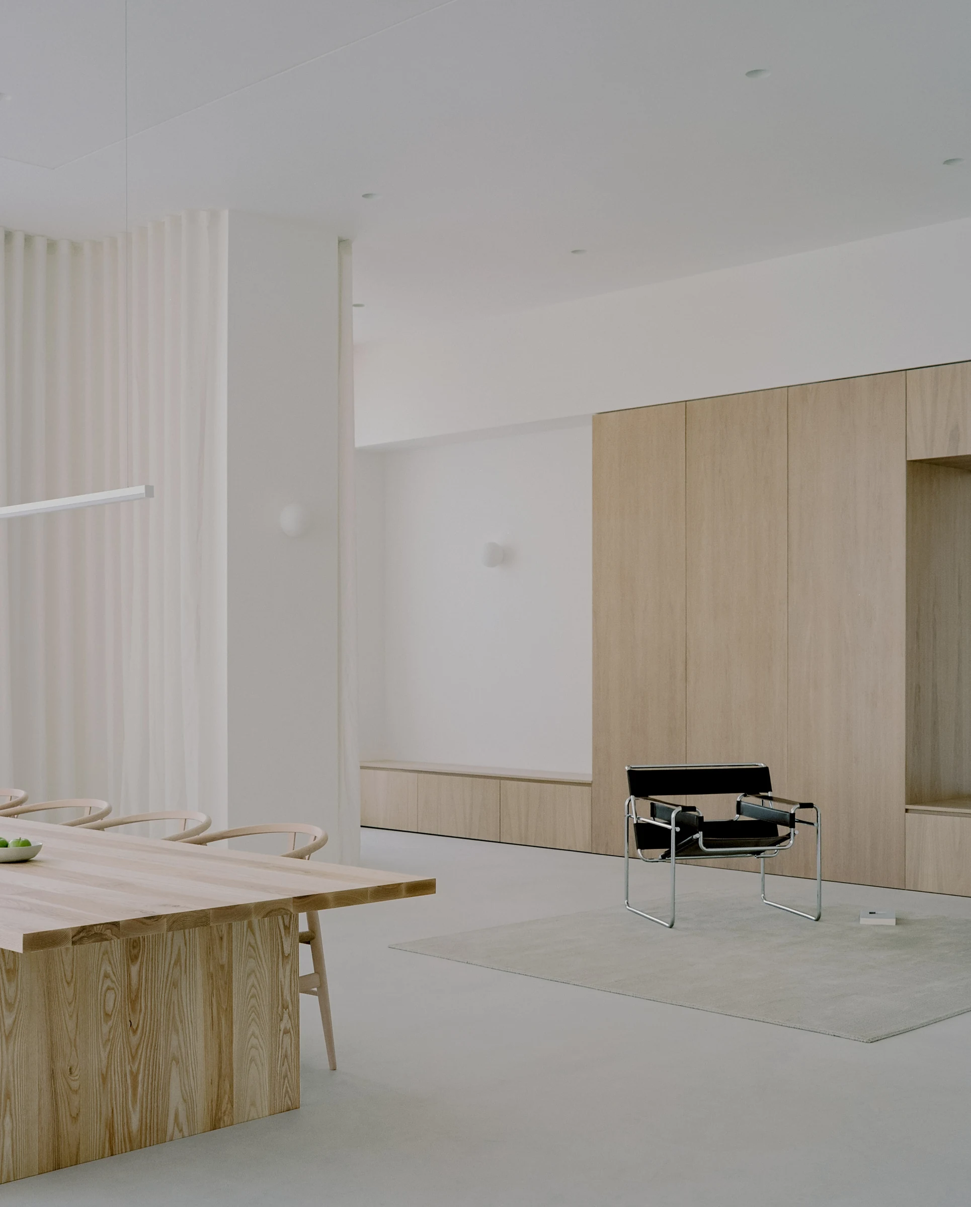 Cherry Orchard by Actual Office, photographed by Nick Dearden — Common room on the first floor: white-oiled oak storage wall at right with a Knoll Wassily chair set on a wool rug, a sheer voile curtain falling at left, a long ash communal table at the…