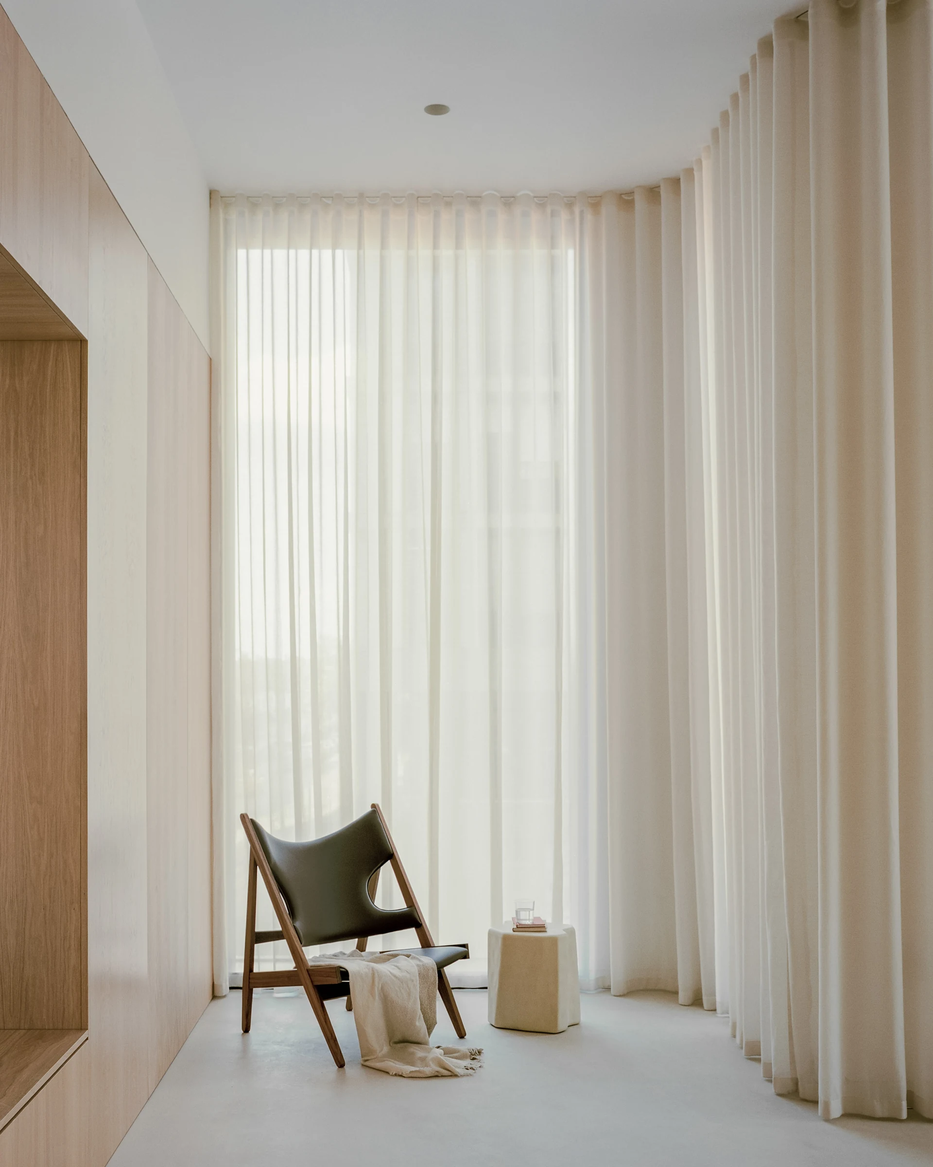 Cherry Orchard by Actual Office, photographed by Nick Dearden — Wider room view: oak joinery wall holding a wall-mounted dark screen and a dark green wool sofa, a breccia stone plinth used as a side table at left, a long ash table with wishbone chairs…