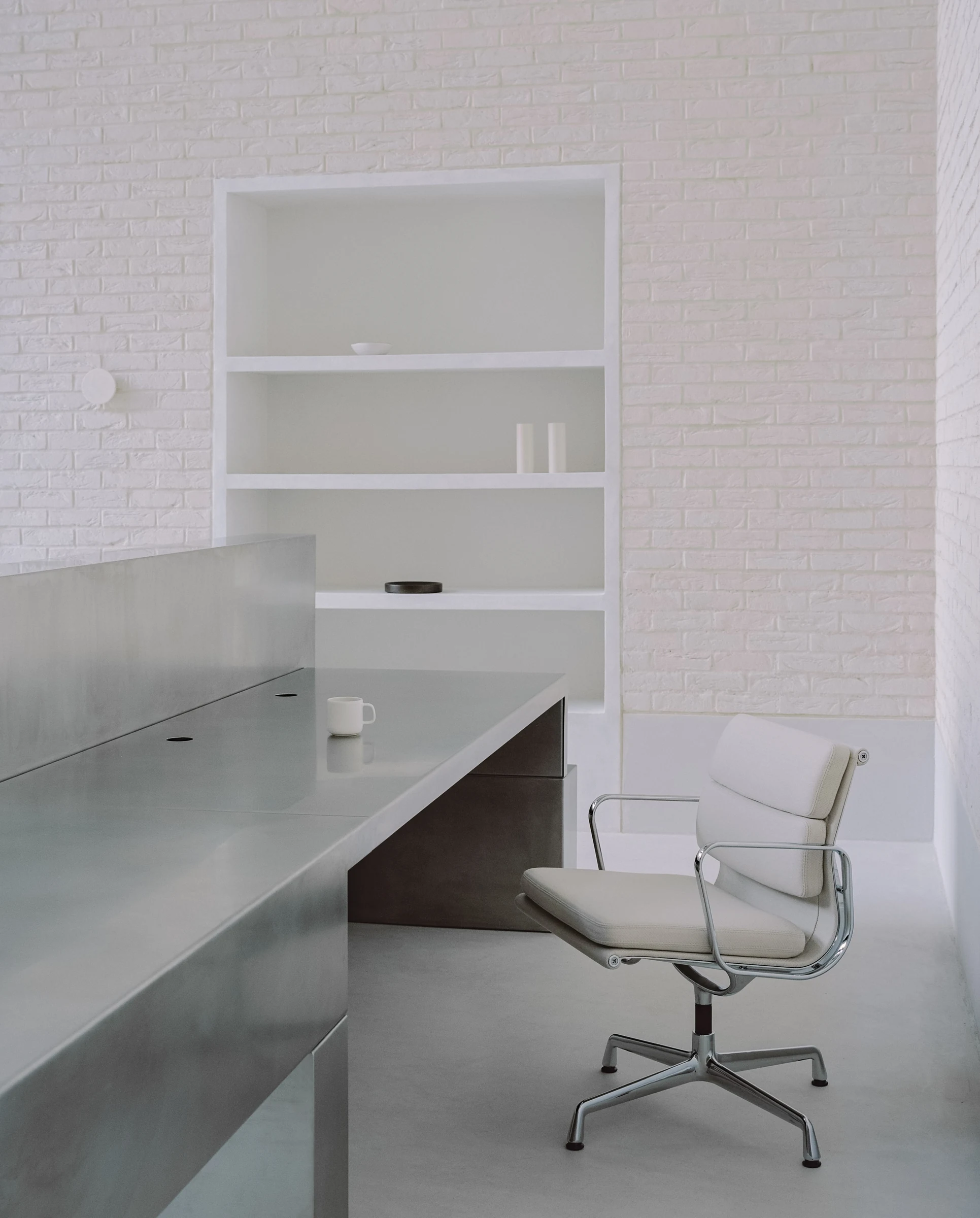 Cherry Orchard by Actual Office, photographed by Nick Dearden — Close view of the concierge desk with a white Eames soft-pad office chair behind it: the stainless surface, a single porcelain cup, the recessed niche shelf in the brick beyond holding two…