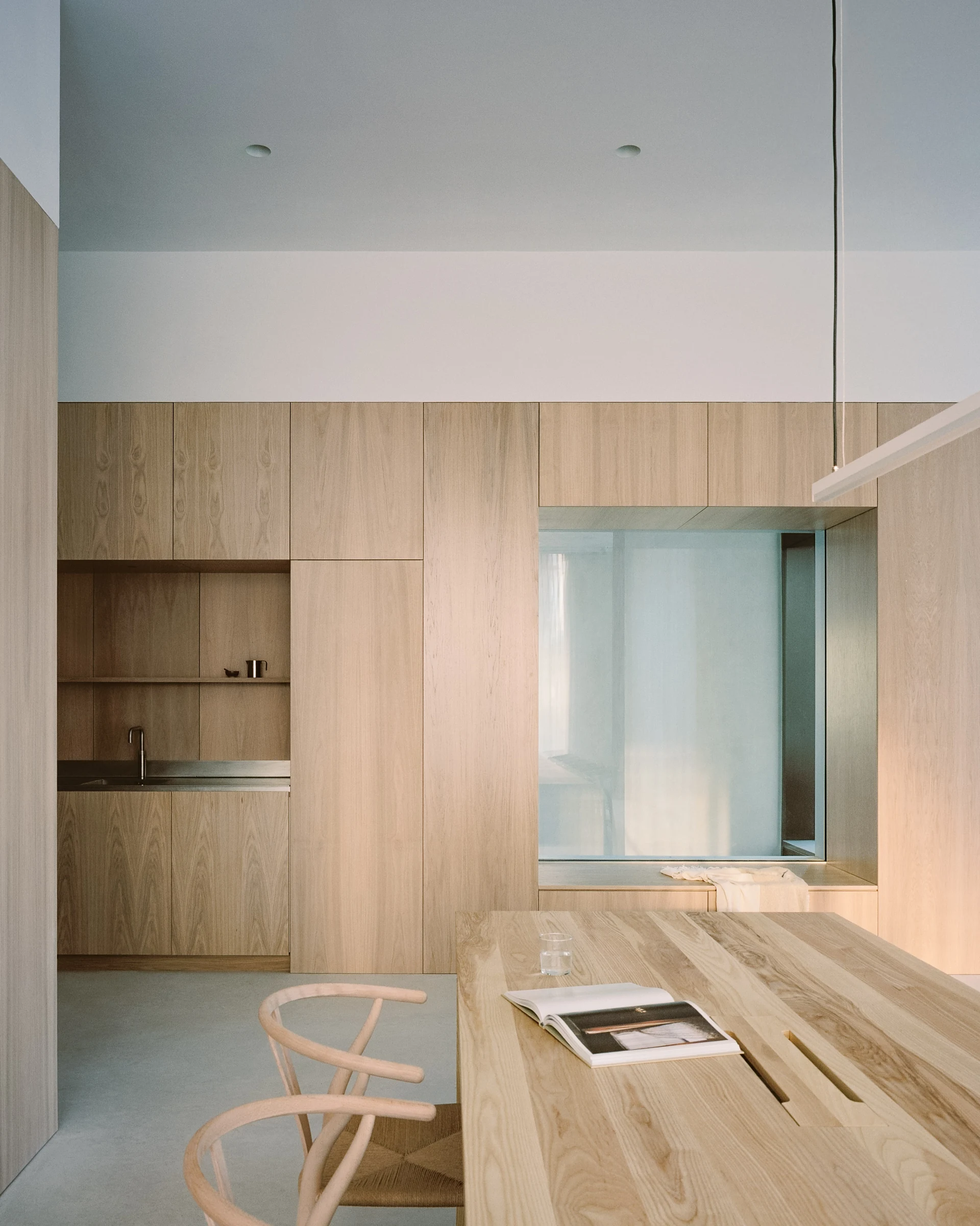 Cherry Orchard by Actual Office, photographed by Nick Dearden — A figure stands at the window wall behind floor-to-ceiling cream voiles, the south London skyline showing through as a pale, low contrast silhouette; long ash communal table and a row of…