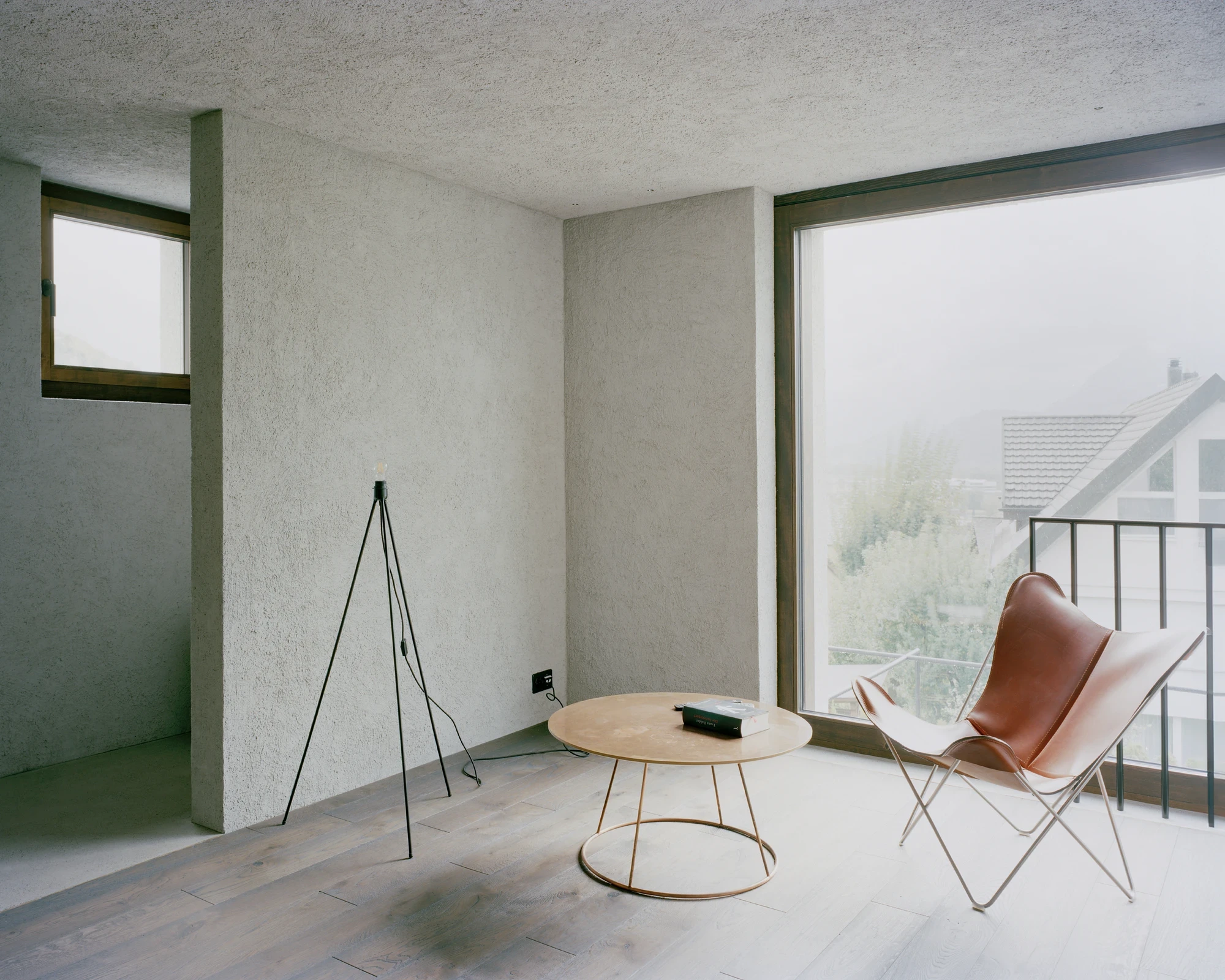Tower house Vaduz by Schreiber Architekten, photographed by Barbara Buehler — a tan leather butterfly chair and a round table sit in a plastered room open to a corner of valley glass.