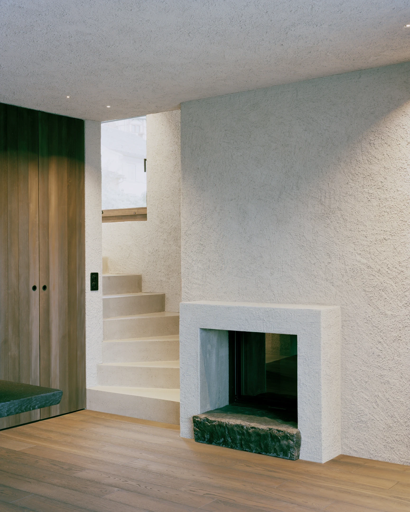 Tower house Vaduz by Schreiber Architekten, photographed by Barbara Buehler — a cast lime fireplace on a rough green stone hearth meets a pale stair and a spruce-clad wall.