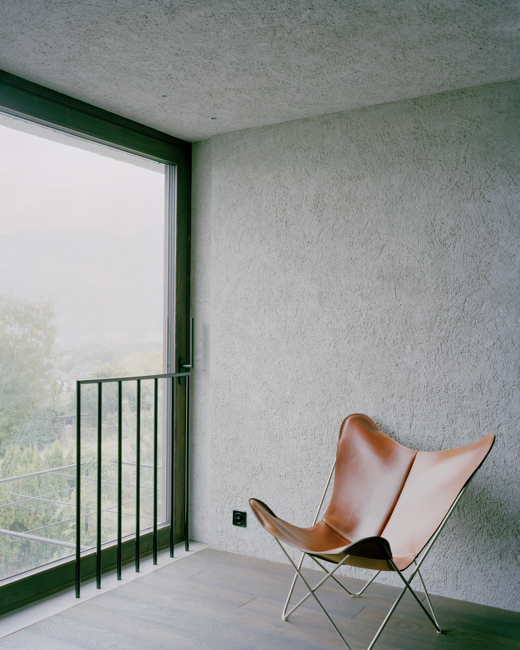 Tower house Vaduz by Schreiber Architekten, photographed by Barbara Buehler — a leather chair rests against troweled gray plaster beside a green-framed window holding the misted valley.