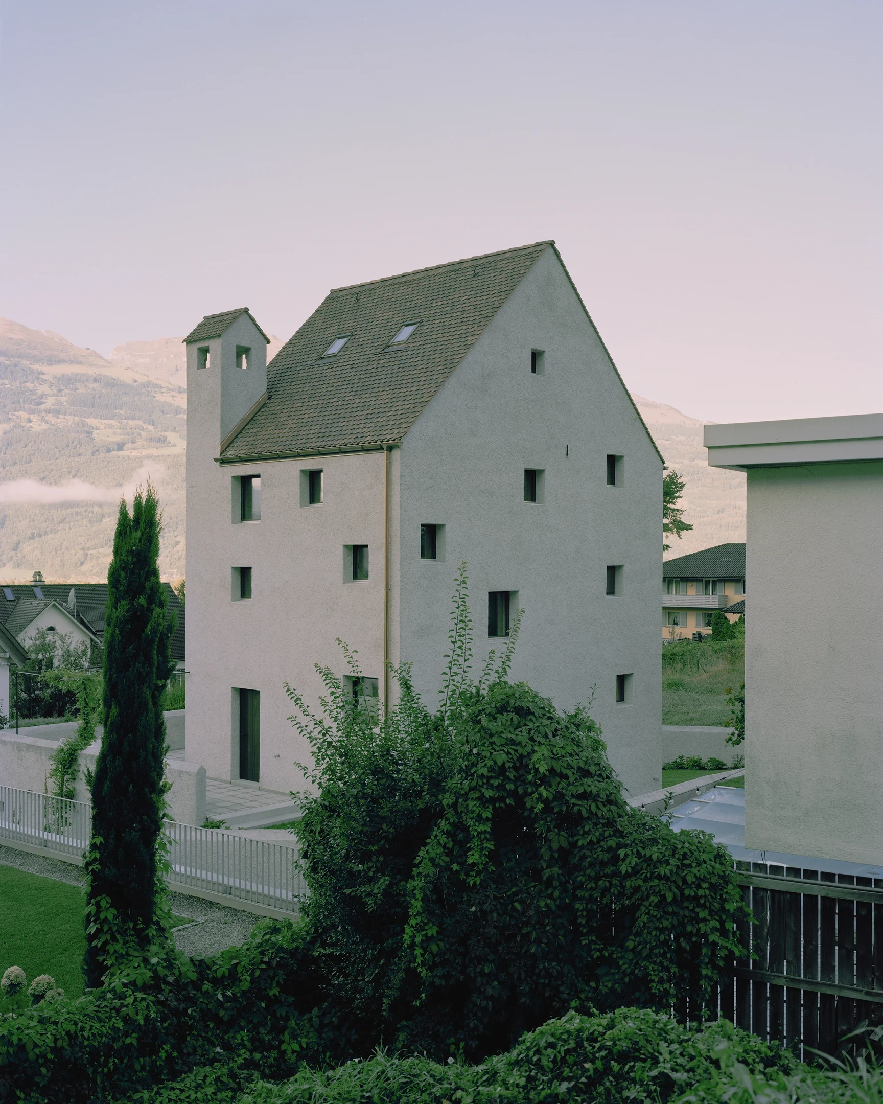 Tower house Vaduz by Schreiber Architekten, photographed by Barbara Buehler — the lime-plastered tower and its tiled gable roof rise above a garden wall against the Alpine slopes at dusk.
