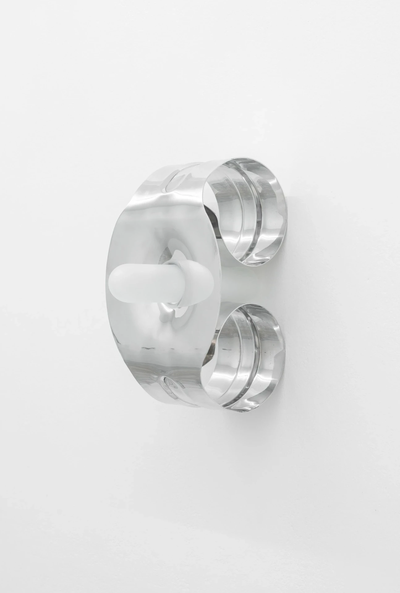 Double Star by Paola Siri Renard, photographed by Dirk Tacke — a mirror-polished double-ring form with twin curled halves, a frosted white bar threaded through its center against the white wall.
