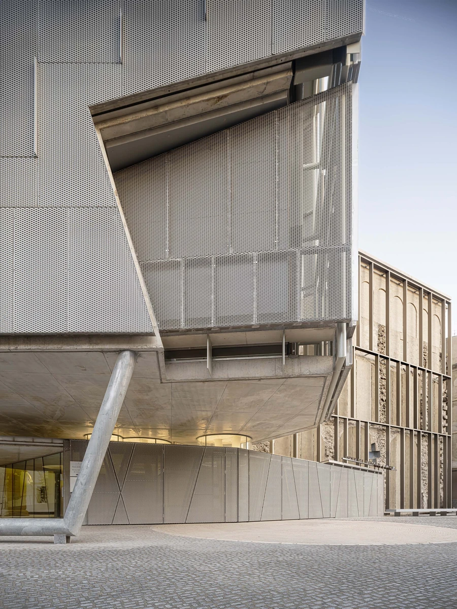 Entrance Façade of the Baroque Museum of Catalonia by David Closes, photographed by Adrià Goula, José Hevia — A perforated-metal volume folds and cantilevers over a glazed entry, the warm old stone wall visible at right in low side light.