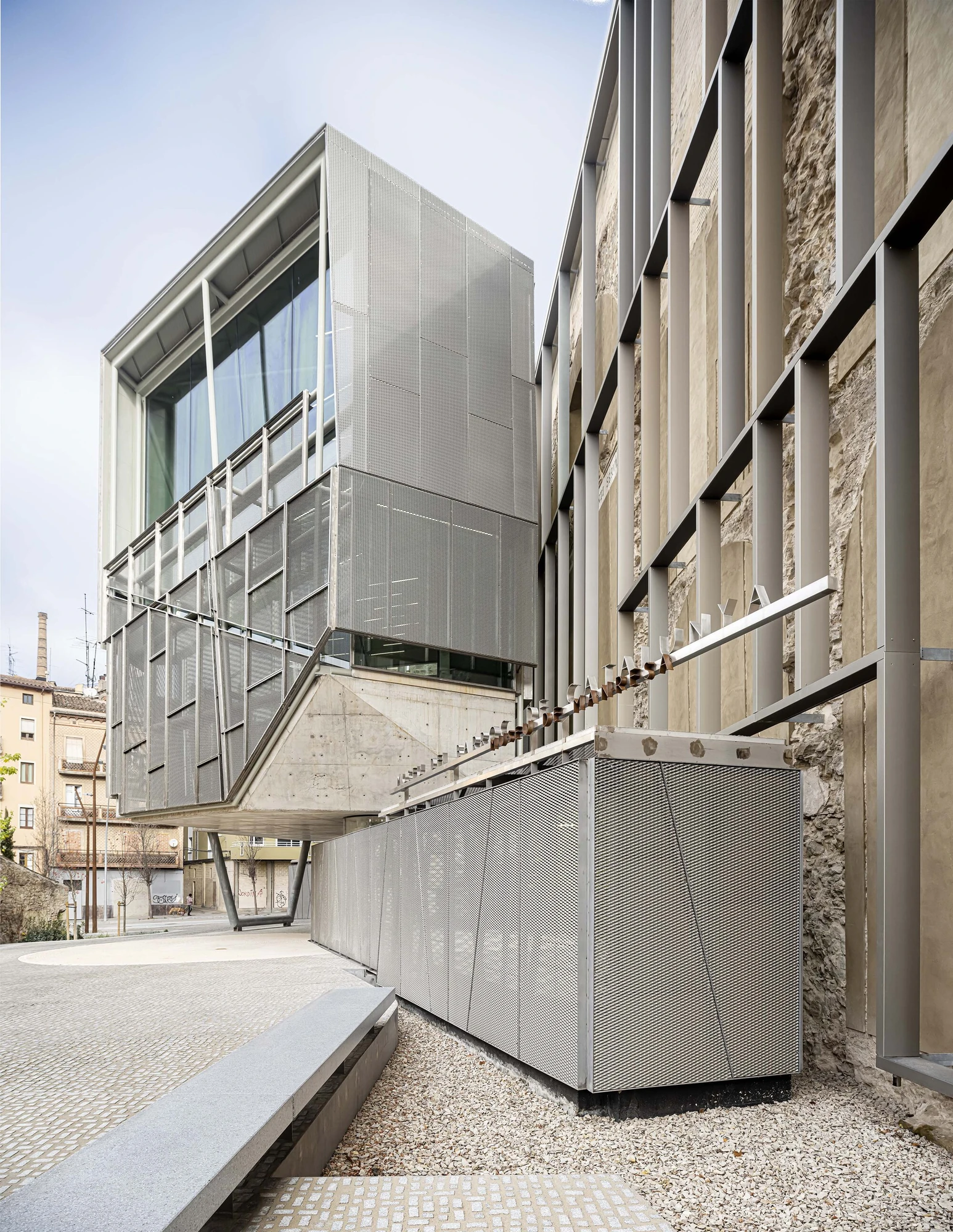 Entrance Façade of the Baroque Museum of Catalonia by David Closes, photographed by Adrià Goula, José Hevia — A glazed upper box rides a cantilever over an expanded-metal counter, gravel and the ghost-arched stone wall running alongside under grey sky.