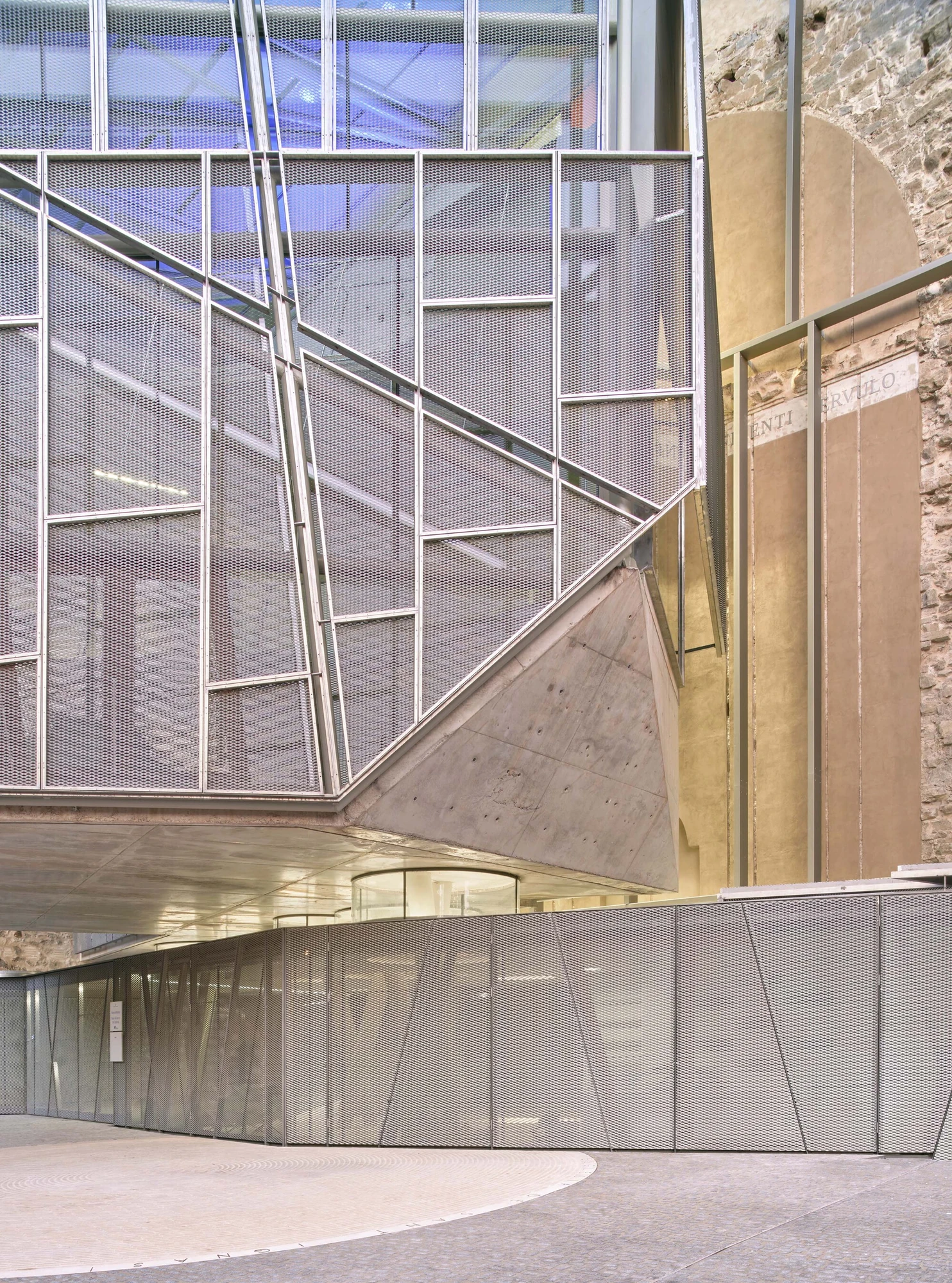 Entrance Façade of the Baroque Museum of Catalonia by David Closes, photographed by Adrià Goula, José Hevia — An atrium where a mesh-clad stair angles past board-formed concrete and an old stone wall still bearing faded painted lettering.