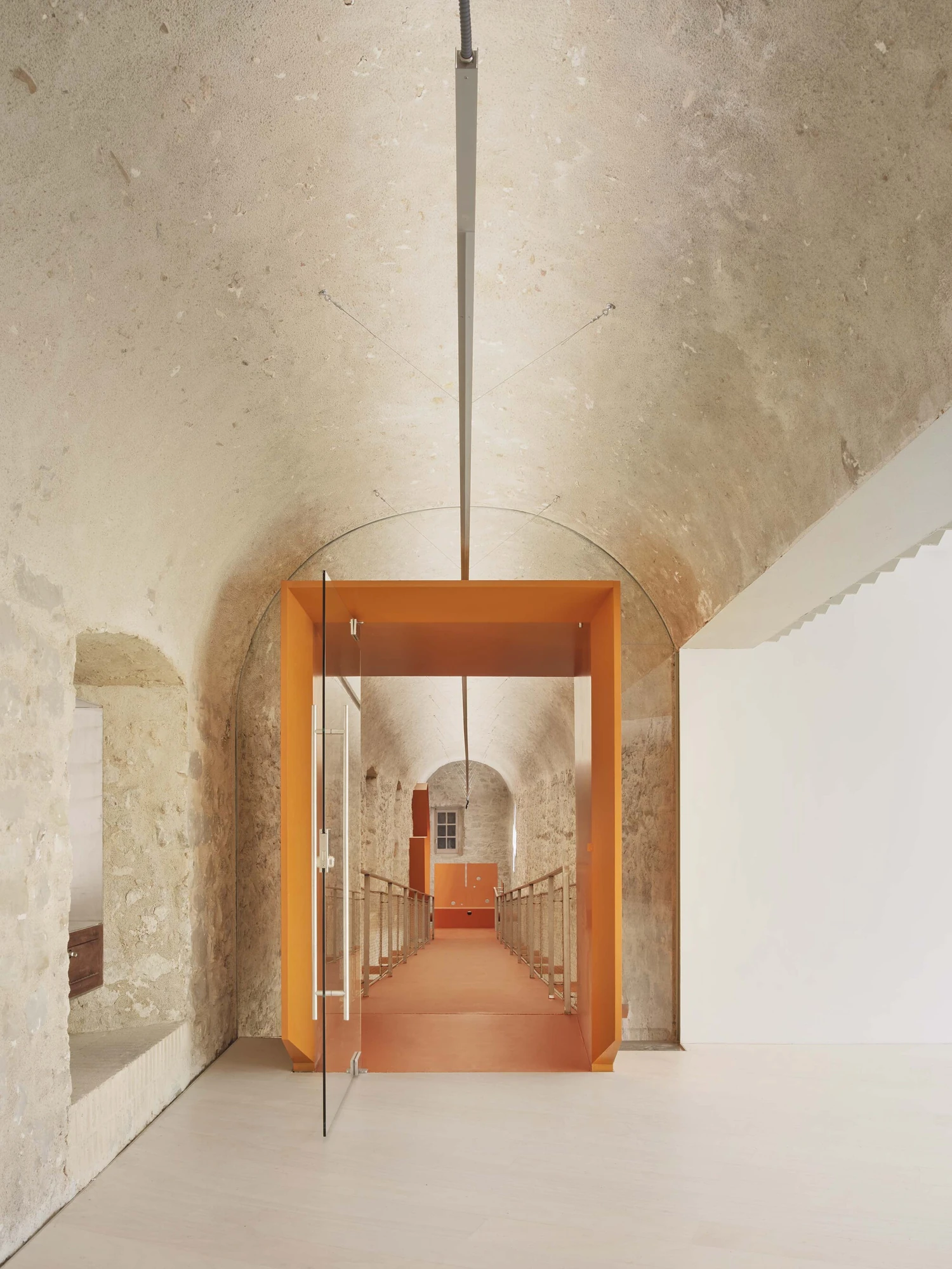 Entrance Façade of the Baroque Museum of Catalonia by David Closes, photographed by Adrià Goula, José Hevia — A white vaulted room opens through a bright orange portal and pivoting glass door onto an orange bridge running deep into the old fabric.