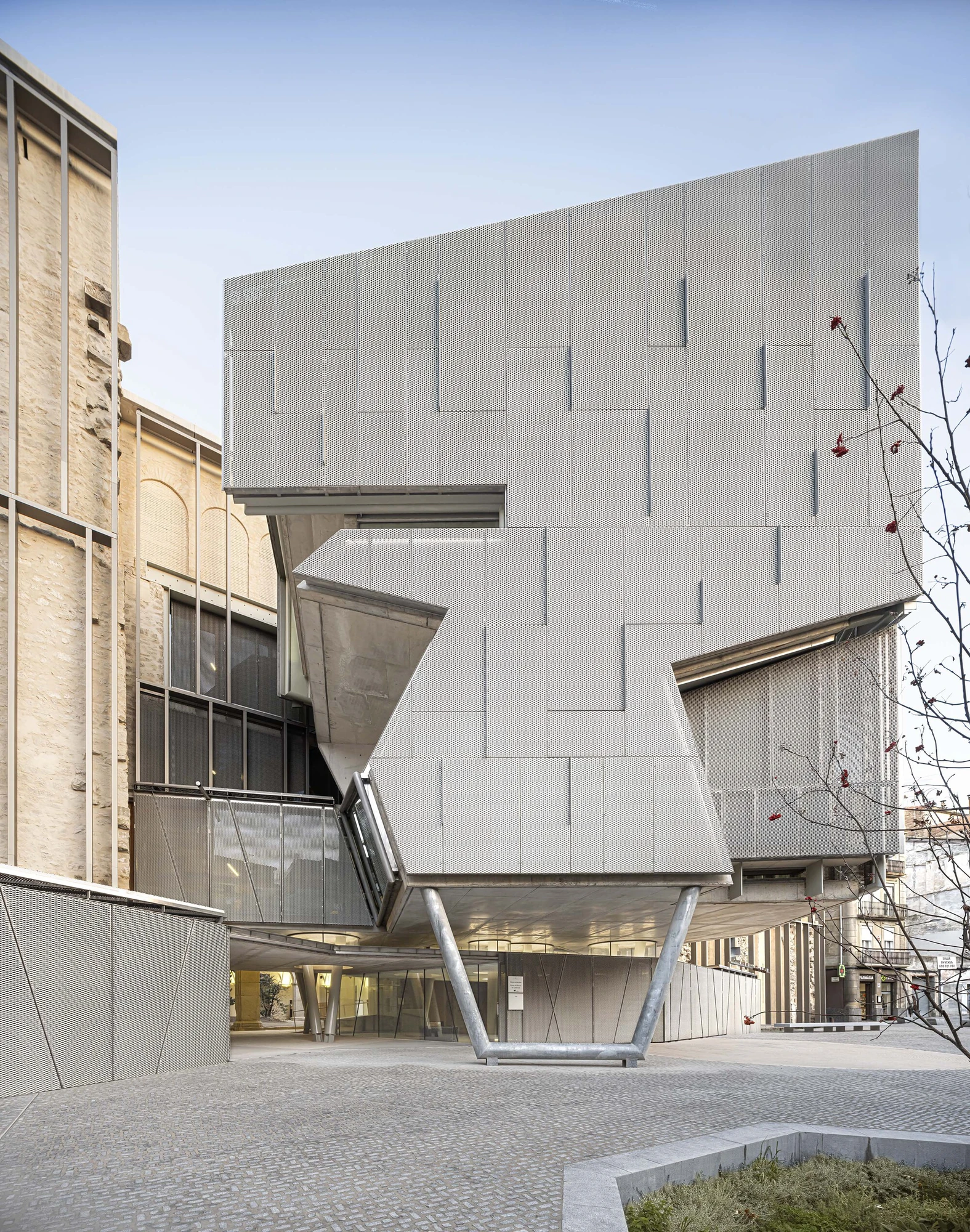 Entrance Façade of the Baroque Museum of Catalonia by David Closes, photographed by Adrià Goula, José Hevia — The perforated-metal mass zigzags in faceted folds on slim steel struts above a glazed ground floor, the scarred church wall flanking it to the…