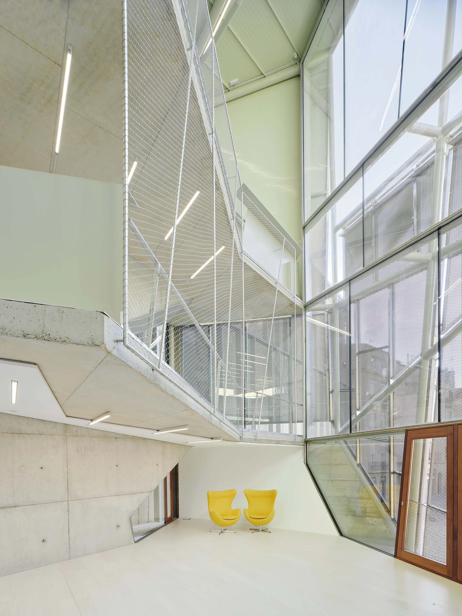 Entrance Façade of the Baroque Museum of Catalonia by David Closes, photographed by Adrià Goula, José Hevia — A tall interior of mesh and angled glazing over board-formed concrete, with two yellow Egg chairs and an oak-framed window set on the pale floor.