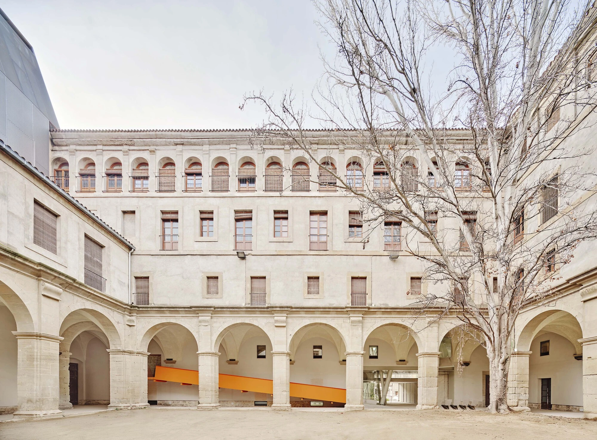 Entrance Façade of the Baroque Museum of Catalonia by David Closes, photographed by Adrià Goula, José Hevia — The arcaded cloister courtyard with stacked stone galleries and a bare winter tree, an orange steel ramp inserted beneath the lower arches.