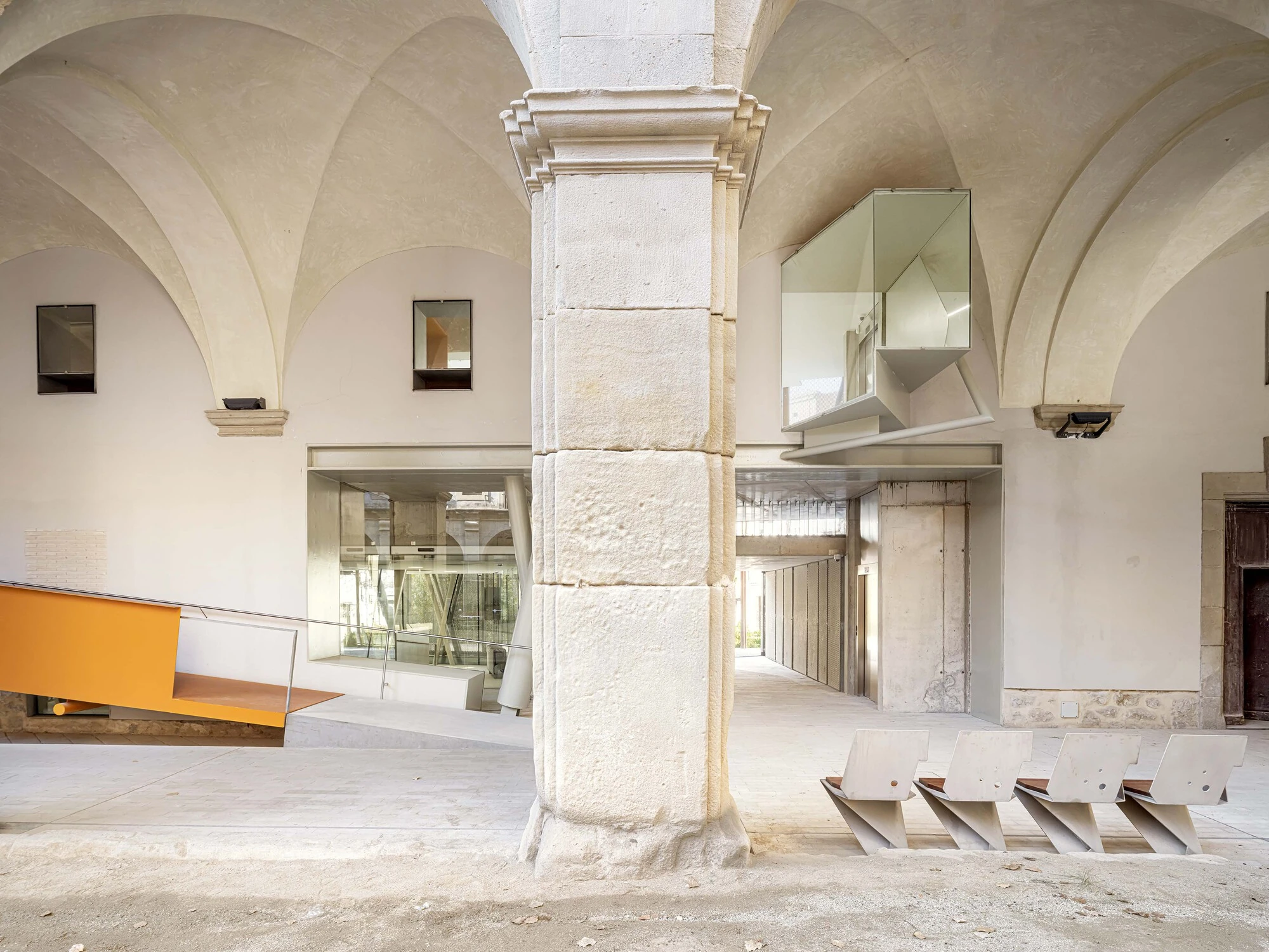 Entrance Façade of the Baroque Museum of Catalonia by David Closes, photographed by Adrià Goula, José Hevia — A massive stone cloister column under groin vaults, a mirrored glass volume cantilevering through one bay while plywood zigzag stools line the…