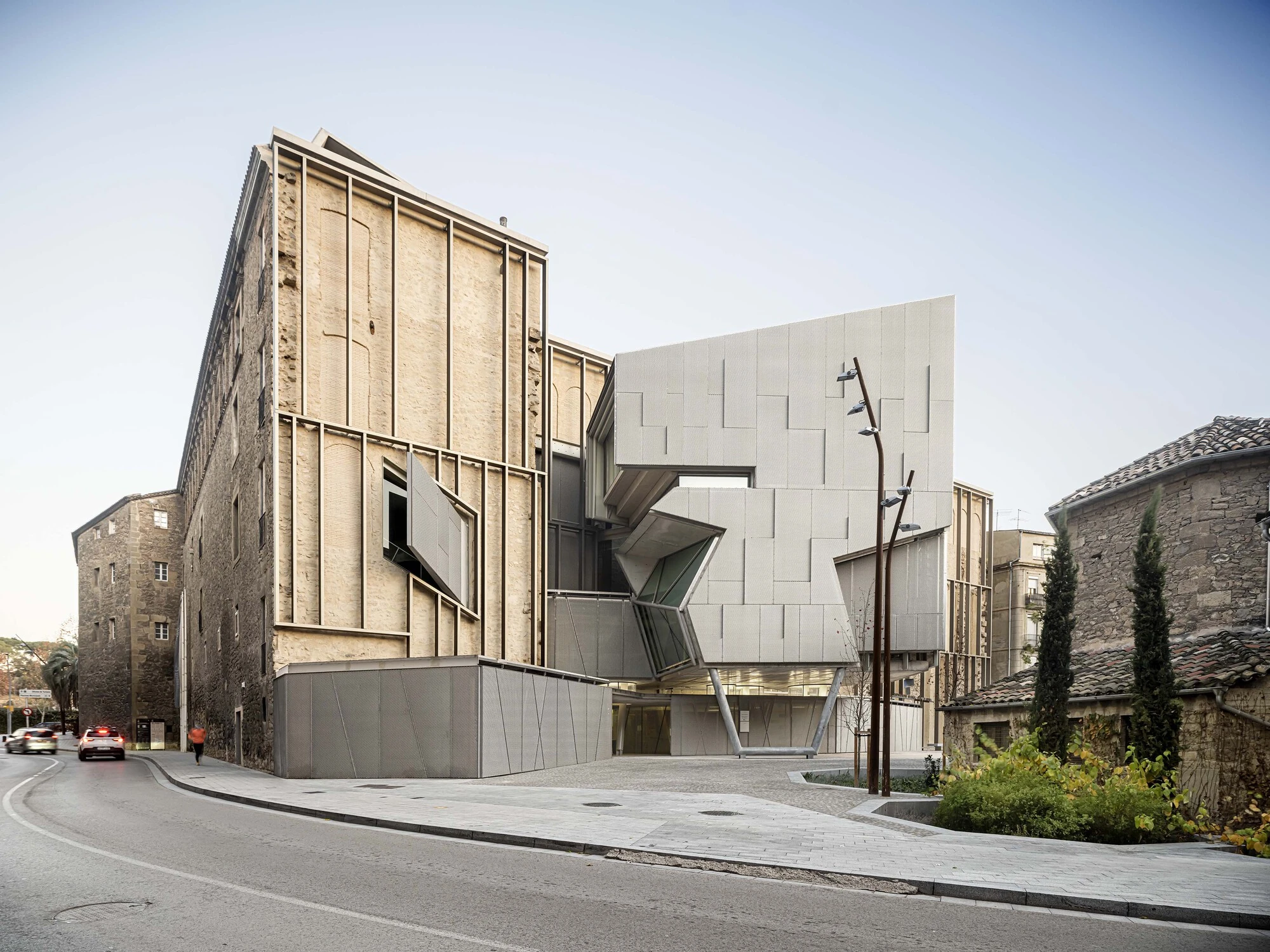 Entrance Façade of the Baroque Museum of Catalonia by David Closes, photographed by Adrià Goula, José Hevia — A wide street view sets the old Jesuit stone wing against the pale silver paneled extension, with cobbled plaza and town rooftops beyond.