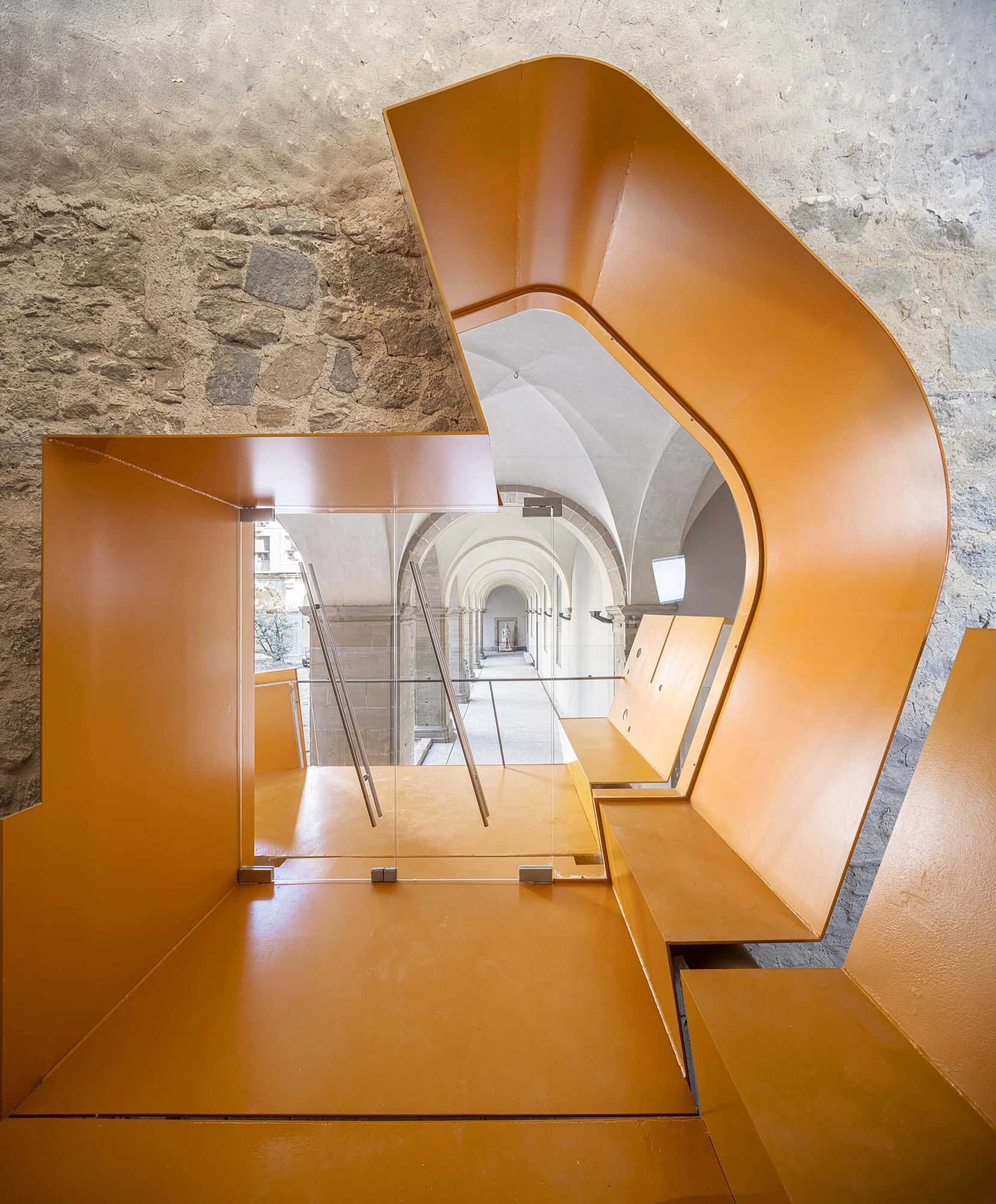 Entrance Façade of the Baroque Museum of Catalonia by David Closes, photographed by Adrià Goula, José Hevia — Folded orange sheet-metal walls and stair fold against rough rubble stone, a glass balustrade catching the vaulted cloister beyond.
