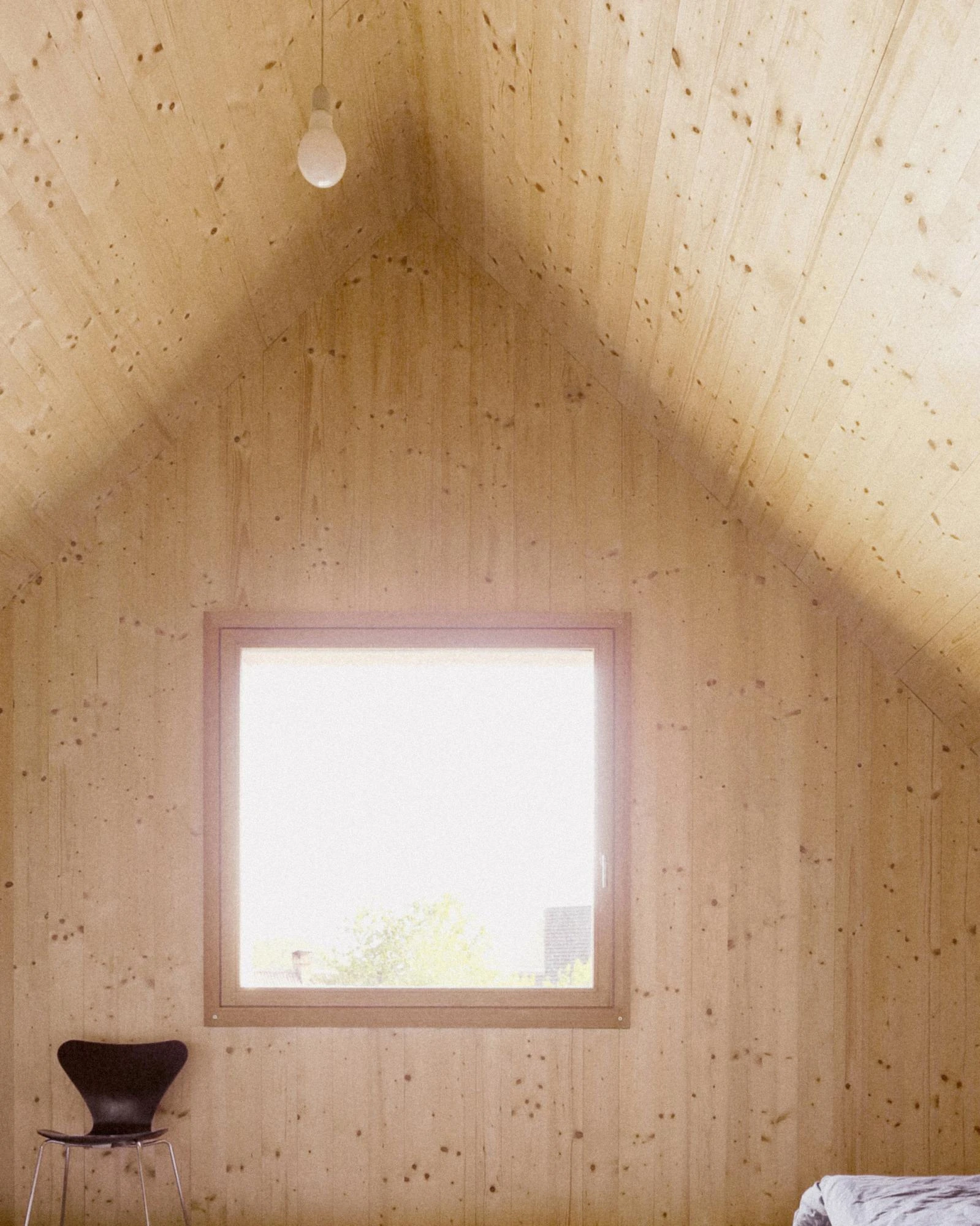 House Alva by Arhitekti Počivašek Petranovič, photographed by Urban Petranovič — A spruce-lined bedroom under the gable, a square window between pitched boards with a black Series 7 chair below.