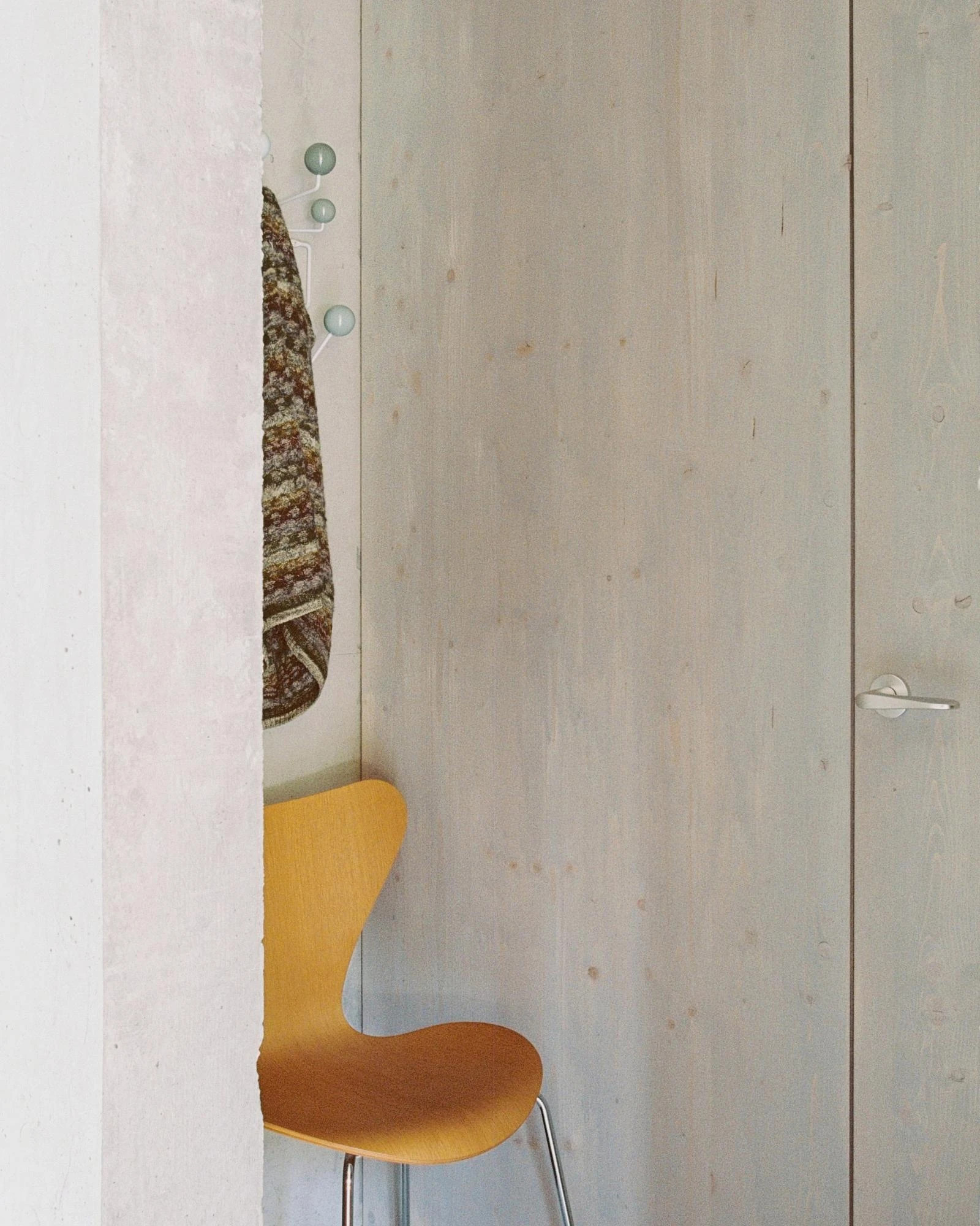 House Alva by Arhitekti Počivašek Petranovič, photographed by Urban Petranovič — A spruce-panelled wall with a yellow Series 7 chair, wall hooks, and a knitted garment hung beside a concrete pier.