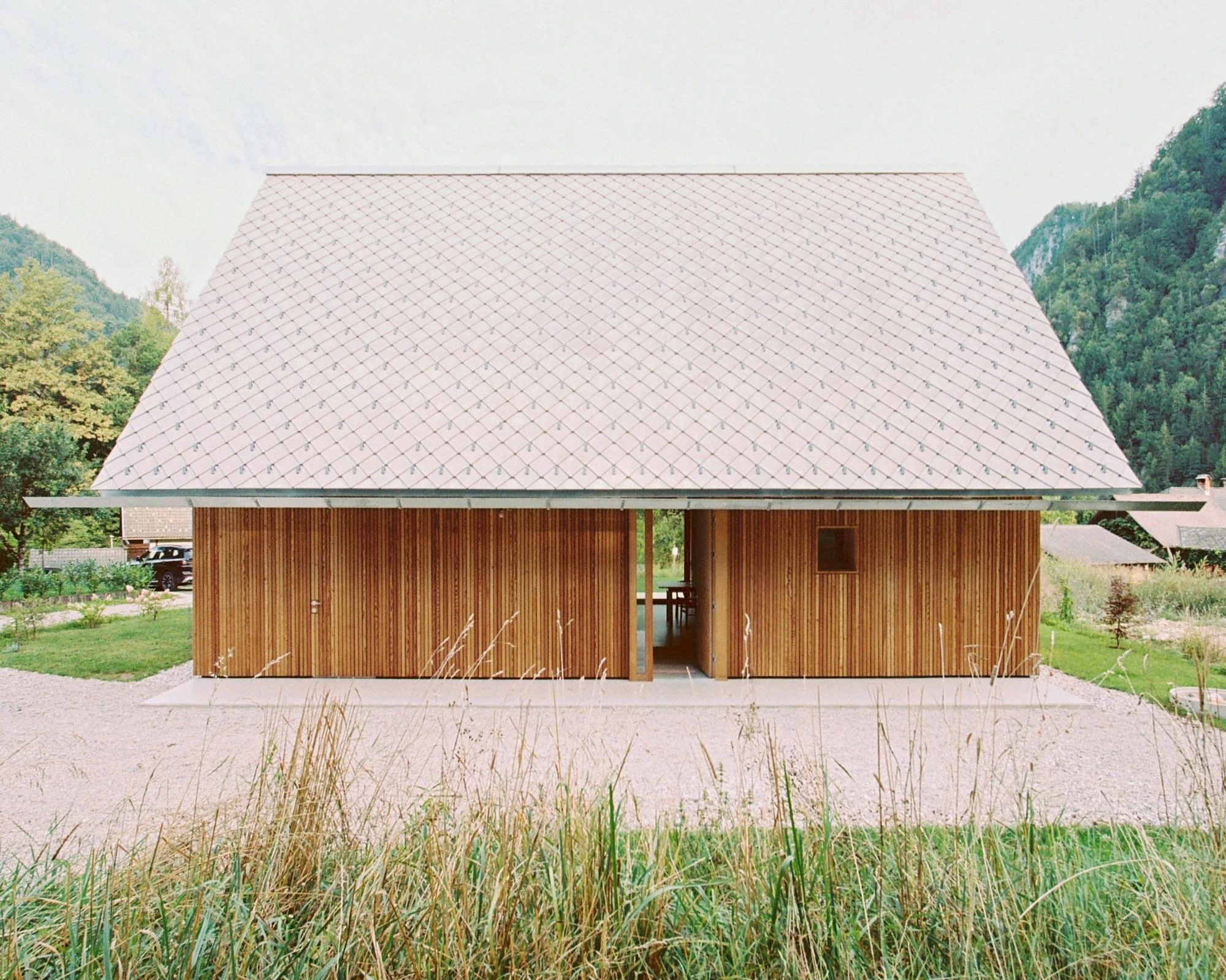 House Alva by Arhitekti Počivašek Petranovič, photographed by Urban Petranovič — The full front of the house in its valley, larch screens drawn back to reveal a through-view across the terrace, mountains behind.