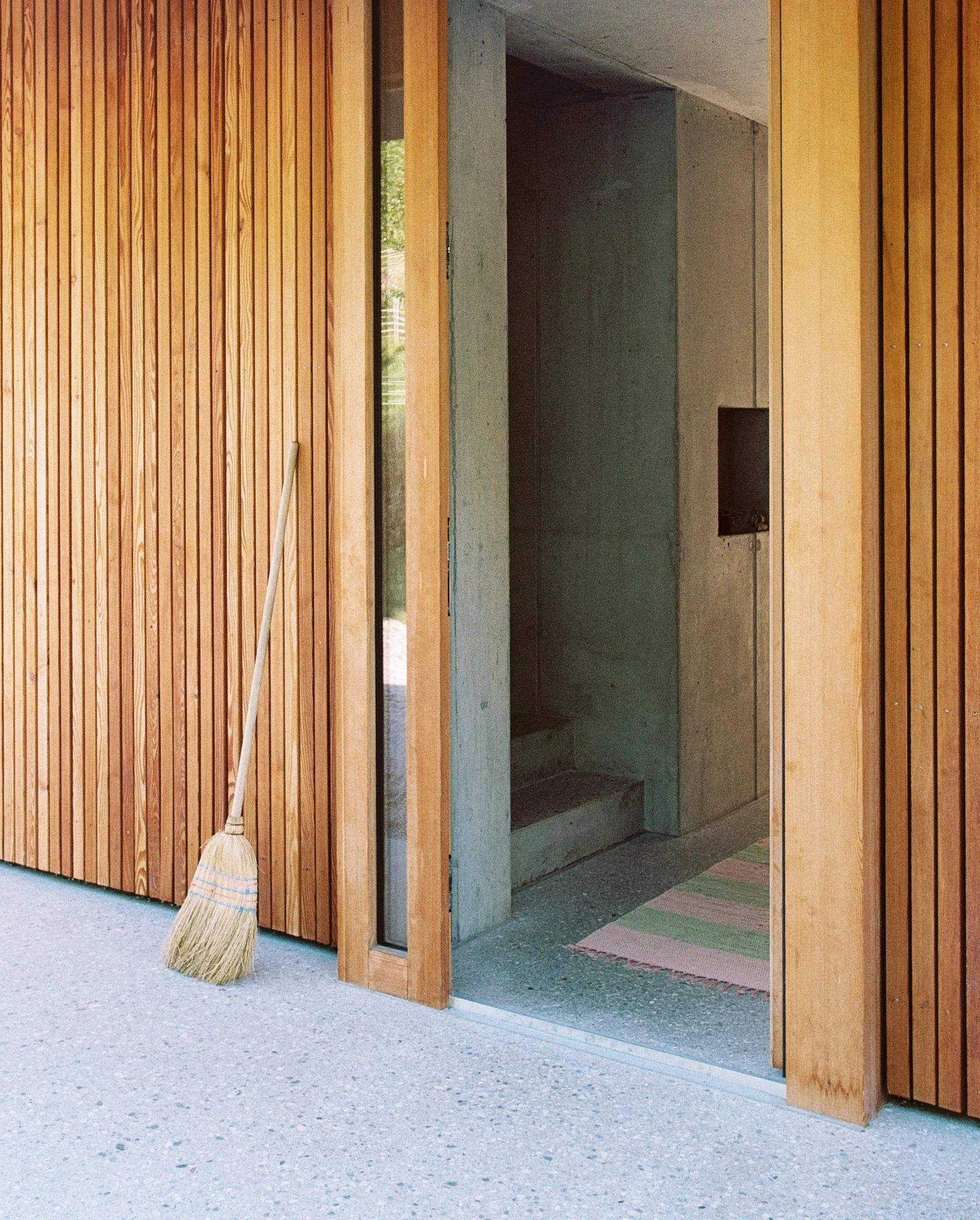 House Alva by Arhitekti Počivašek Petranovič, photographed by Urban Petranovič — An entrance detail of larch slats and a glass slot opening onto a raw concrete hall, a straw broom leaning at the threshold.