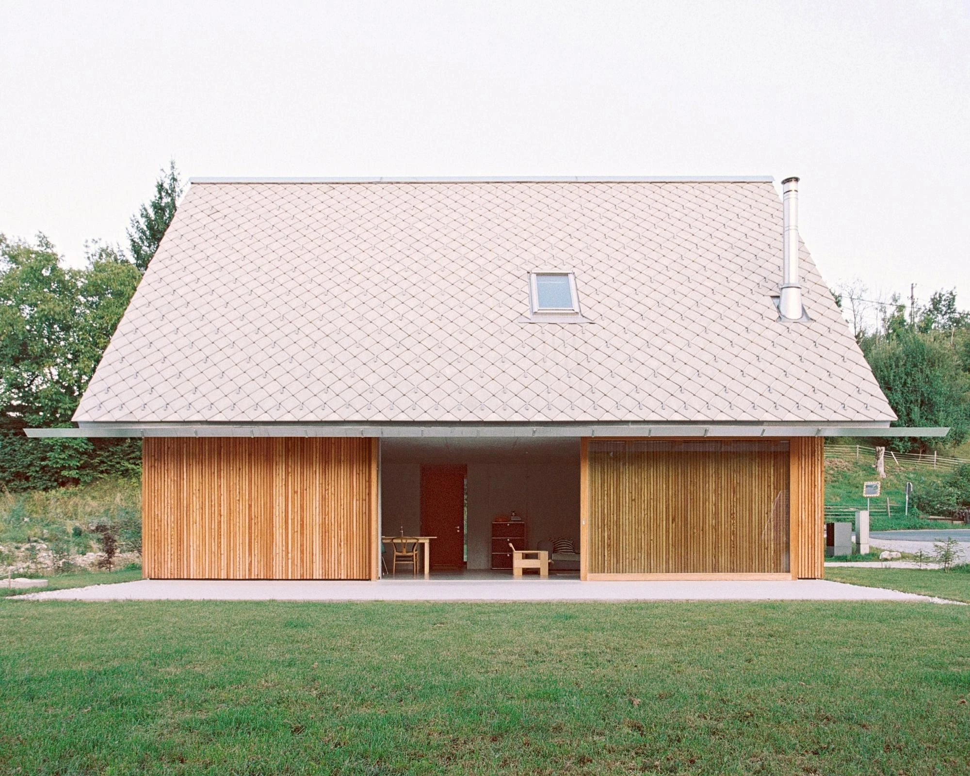 House Alva by Arhitekti Počivašek Petranovič, photographed by Urban Petranovič — The gabled house seen head-on, its diamond-patterned concrete-tile roof sweeping low over open larch screens and a recessed terrace.