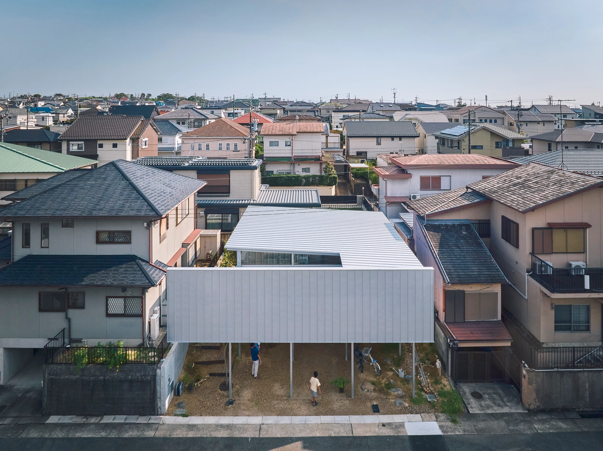 House in Shinokaze by Ikehara Architect & Associates, photographed by Takuya Seki — An elevated view sets the long silver volume among the pitched tiled roofs of the neighborhood, with people moving in the gravel undercroft below.