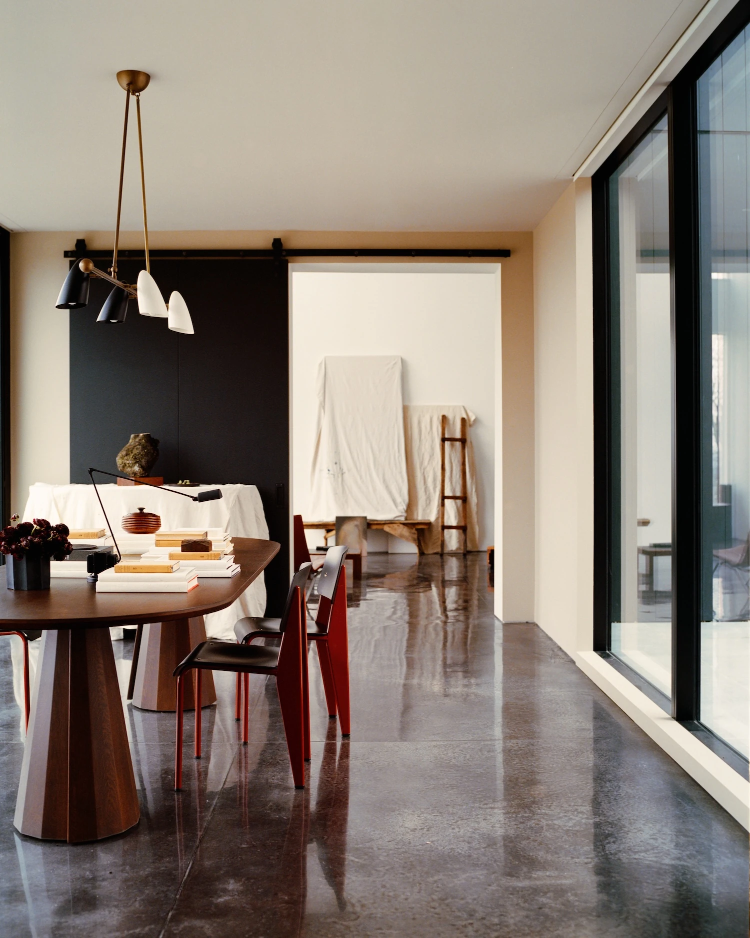 Schoolhouse by JG Neukomm Architecture, photographed by William Jess Laird — A black sliding barn door and brass pendant frame a walnut dining table with red and black chairs reflected in the wet-looking concrete floor.