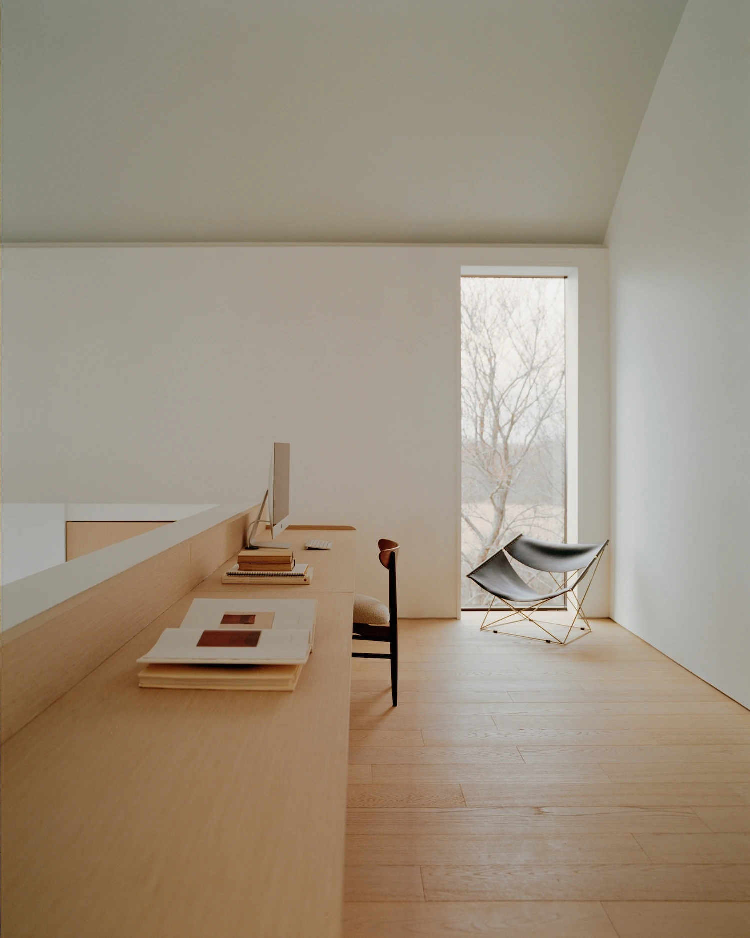 Schoolhouse by JG Neukomm Architecture, photographed by William Jess Laird — A long built-in white oak desk runs the length of a mezzanine office beside a narrow full-height window framing bare winter trees.