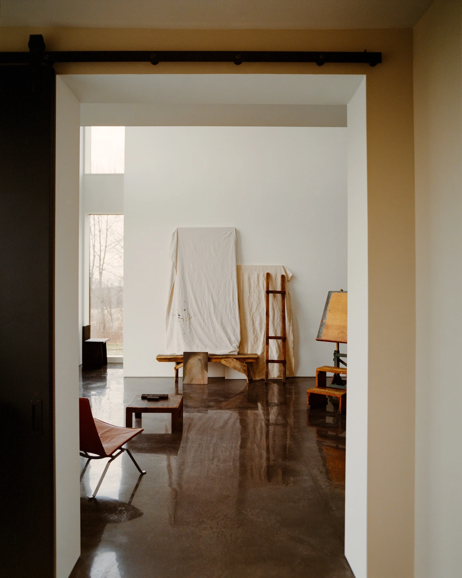 Schoolhouse by JG Neukomm Architecture, photographed by William Jess Laird — Through a dark sliding door, the view opens onto the studio where draped canvases lean against a white wall above mirror-polished concrete.