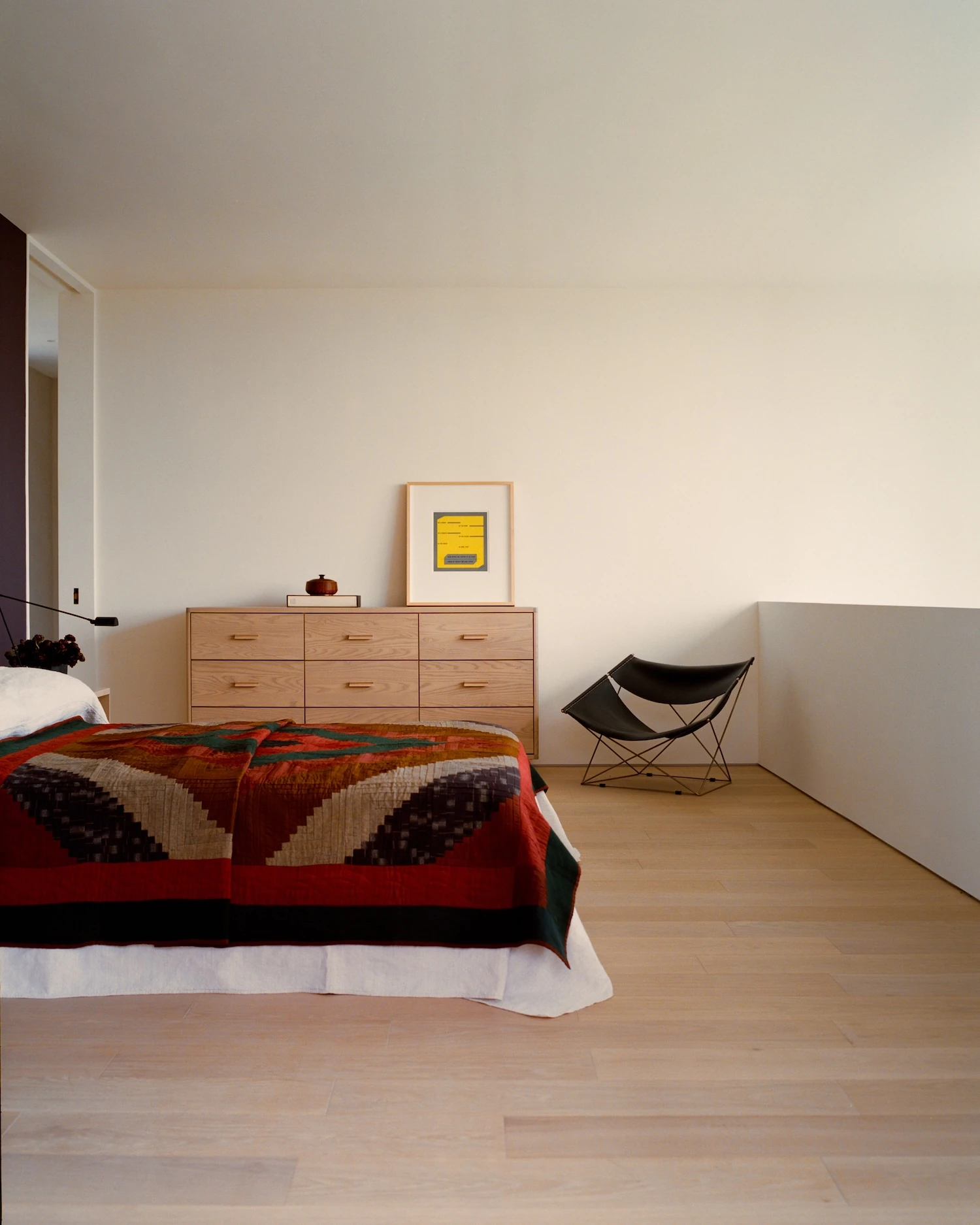 Schoolhouse by JG Neukomm Architecture, photographed by William Jess Laird — In the loft bedroom, an oak dresser and a patchwork quilt sit on pale oak flooring near a black butterfly chair and a yellow-framed print.