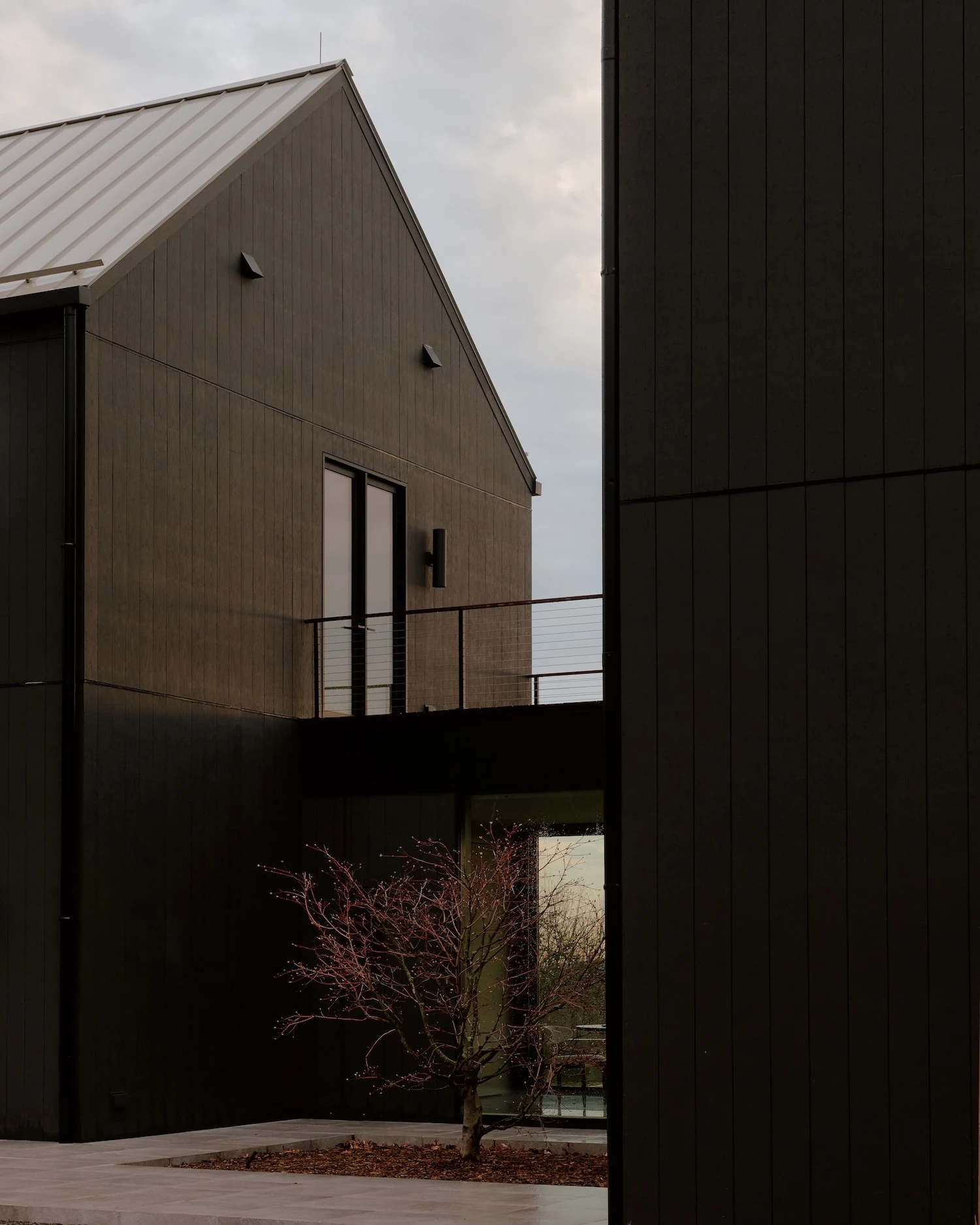 Schoolhouse by JG Neukomm Architecture, photographed by William Jess Laird — At dusk, a narrow gap between dark-clad gabled volumes reveals a cantilevered balcony and a red-budded tree against a grey sky.