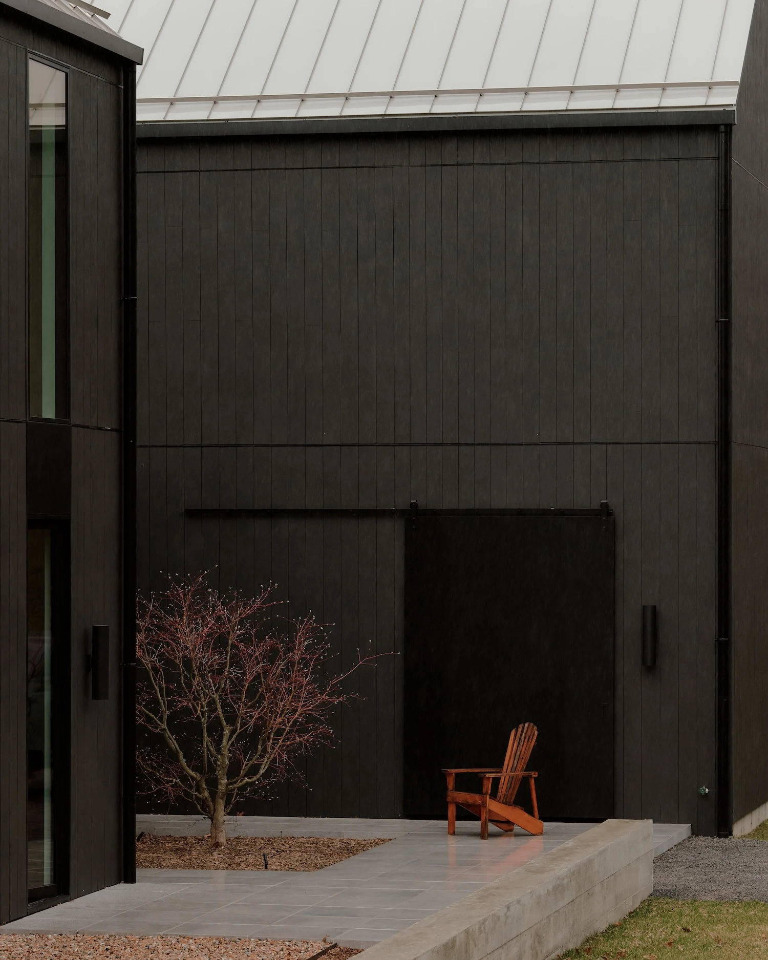 Schoolhouse by JG Neukomm Architecture, photographed by William Jess Laird — A black sliding barn-door panel sits flush in the dark board facade beside a cedar Adirondack chair and a budding Japanese maple on a concrete terrace.