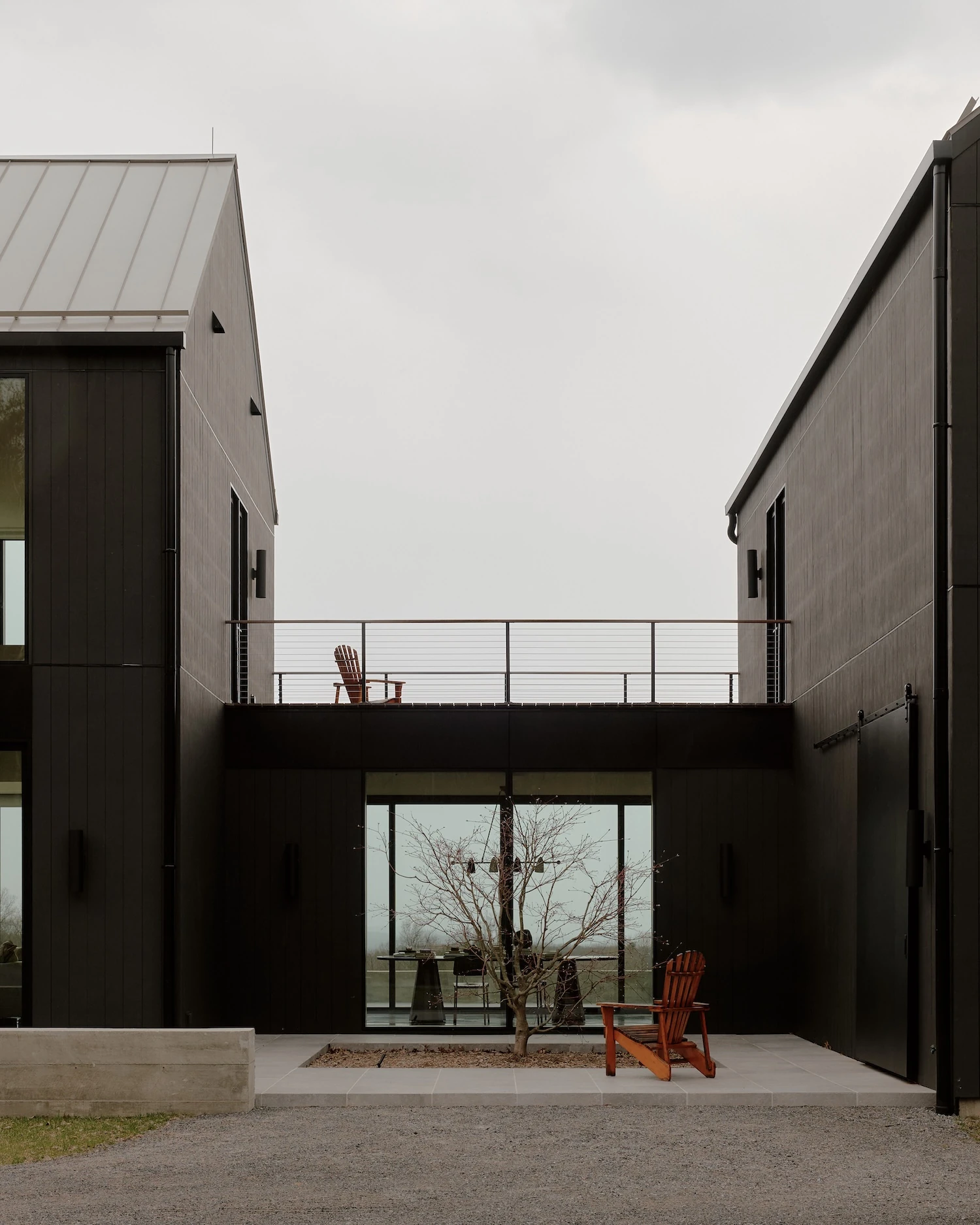 Schoolhouse by JG Neukomm Architecture, photographed by William Jess Laird — The courtyard between the two wings frames a cable-railed rooftop terrace above the glazed gallery, a single bare tree set in the paving below.