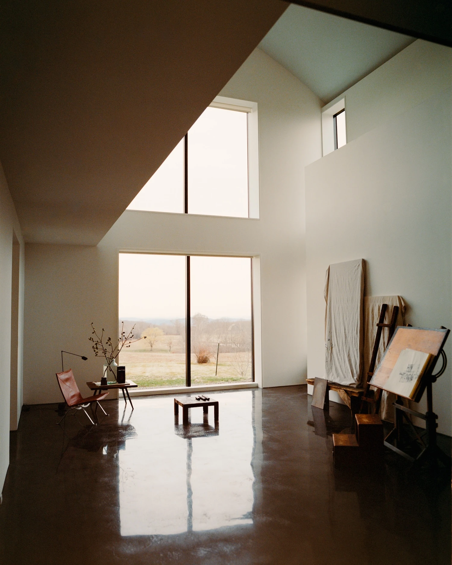 Schoolhouse by JG Neukomm Architecture, photographed by William Jess Laird — Daylight pools across glossy stained concrete in the studio, where wooden easels and a leather sling chair gather below two stacked windows.