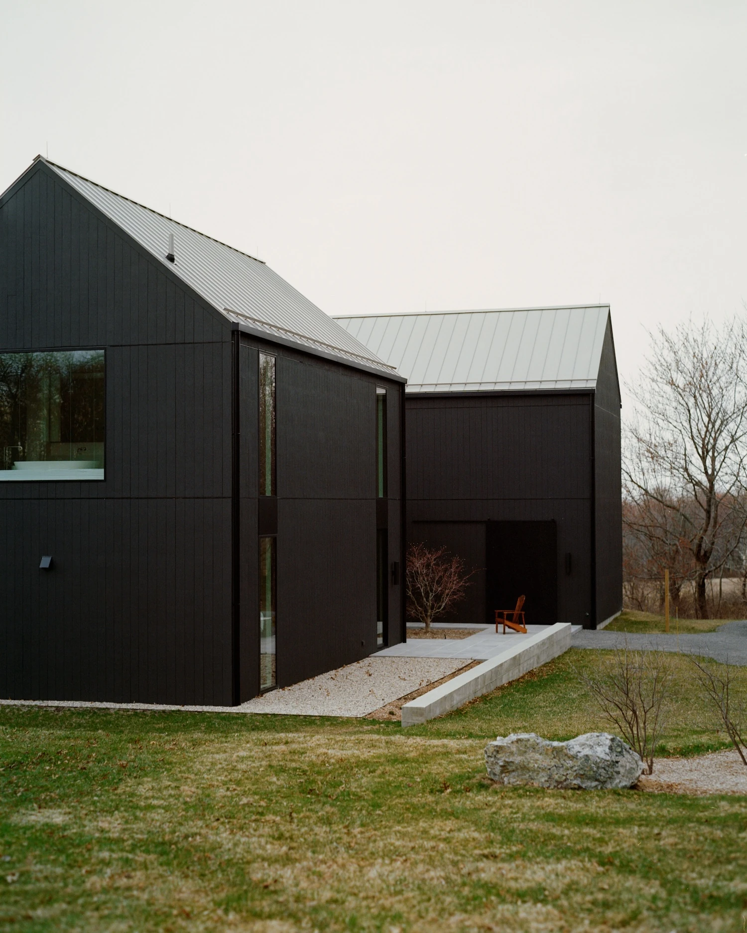 Schoolhouse by JG Neukomm Architecture, photographed by William Jess Laird — Seen across the lawn, the two black gabled volumes under standing-seam metal roofs step down the hill past a concrete ramp and a lone boulder.
