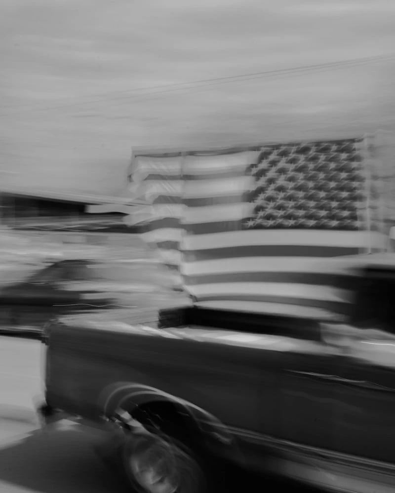 It's Not the Wind by Jordan Conway, photographed by Jordan Conway — A pickup truck carrying an oversized American flag streaks past in motion-blurred black and white, the stripes smeared into pure speed.