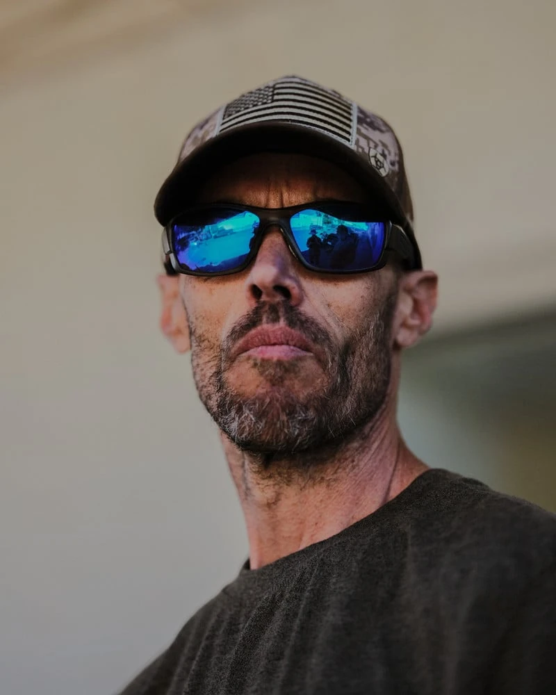 It's Not the Wind by Jordan Conway, photographed by Jordan Conway — A weathered man in a flag-print trucker cap stares down the lens, the photographer and a small crowd reflected in his mirrored blue sunglasses.