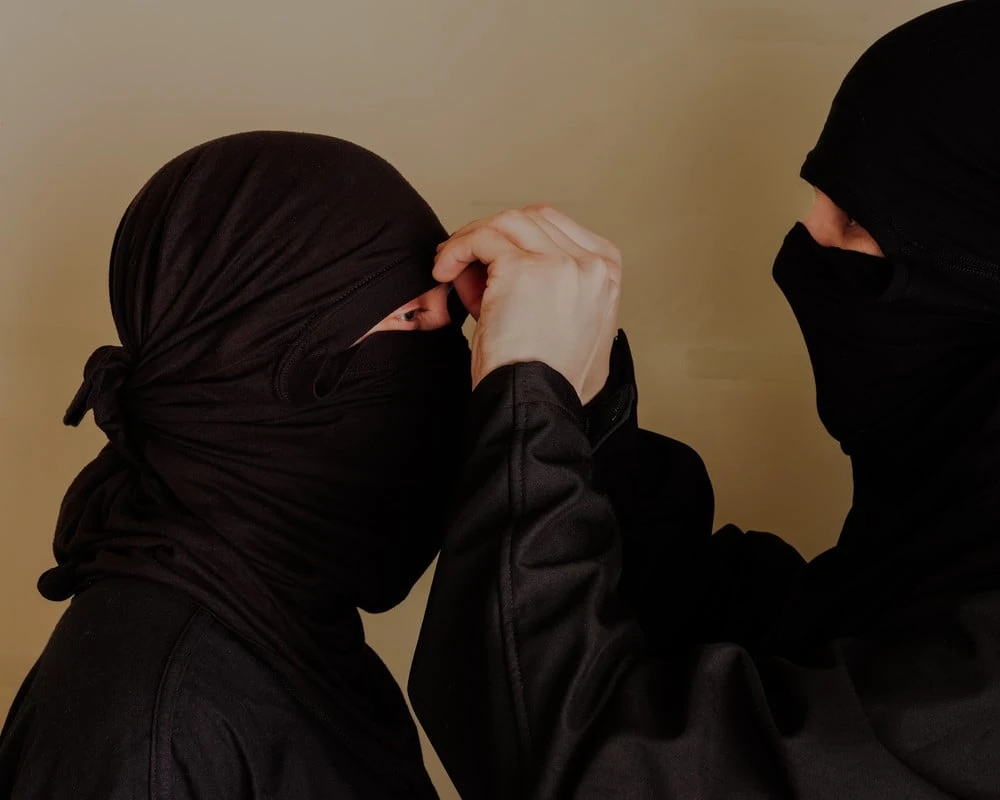 It's Not the Wind by Jordan Conway, photographed by Jordan Conway — Two people in black balaclavas against a beige wall, one reaching across to adjust the fabric over the other's eyes.