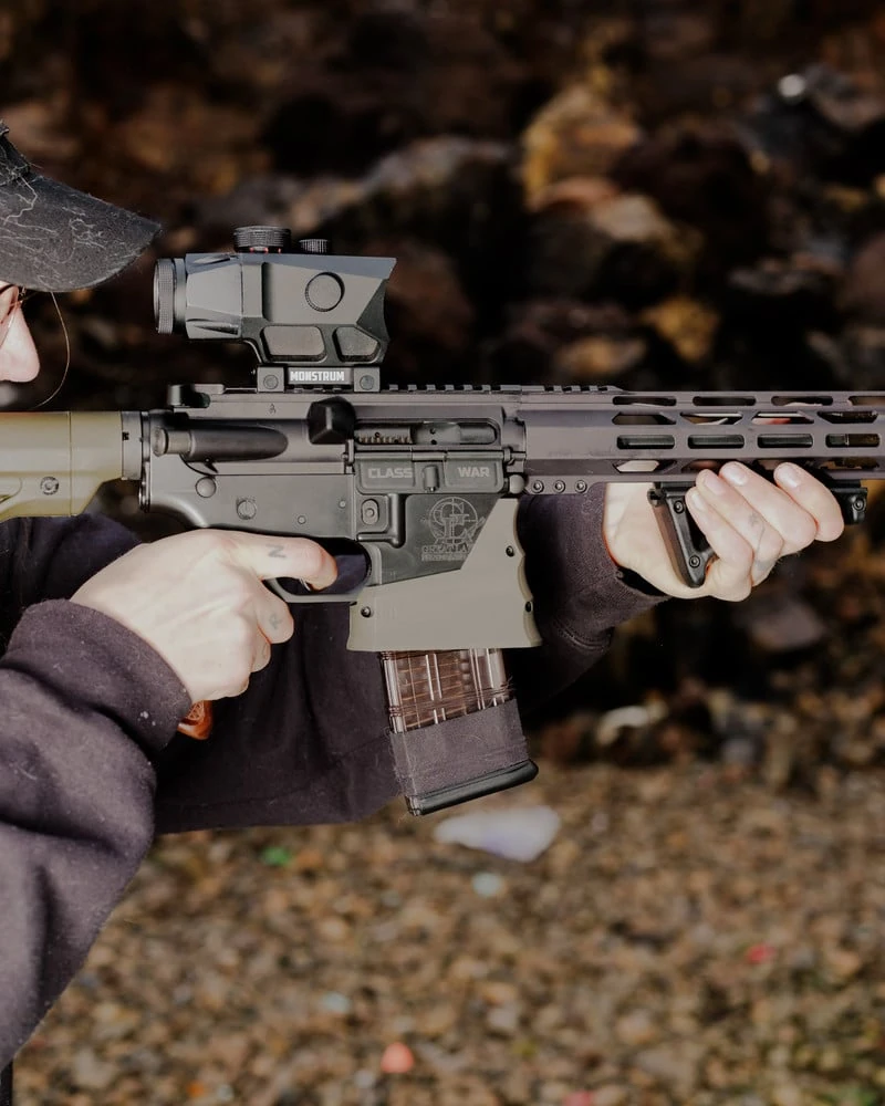 It's Not the Wind by Jordan Conway, photographed by Jordan Conway — Hands cradle an AR-15 rifle whose lower receiver is stamped CLASS WAR, the optic and clear magazine sharp against a blurred rocky ground.