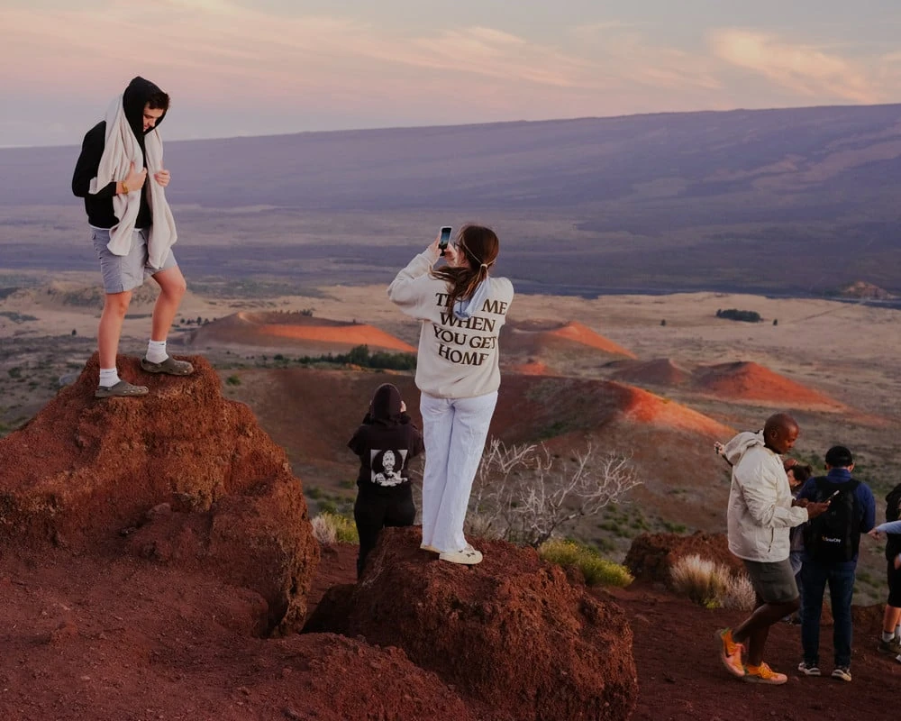 It's Not the Wind by Jordan Conway, photographed by Jordan Conway — Tourists climb red volcanic cinder cones at dusk while a woman photographs the view in a sweatshirt reading TEXT ME WHEN YOU GET HOME.