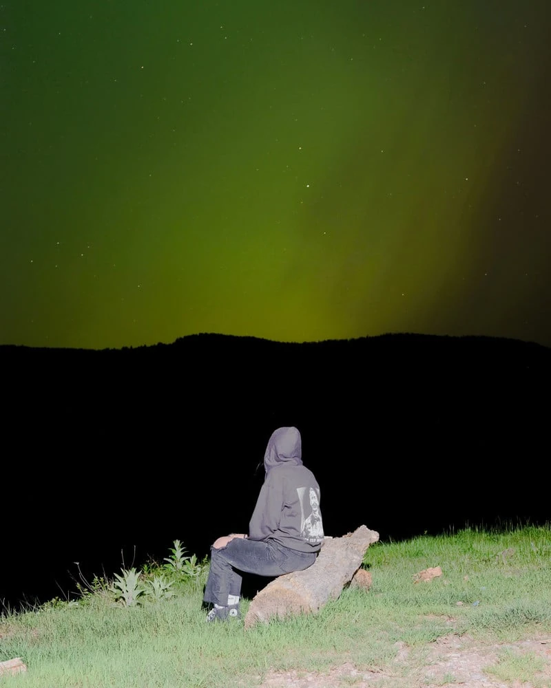 It's Not the Wind by Jordan Conway, photographed by Jordan Conway — A lone hooded figure sits on a fallen log watching a green aurora flood the night sky over a black ridgeline.