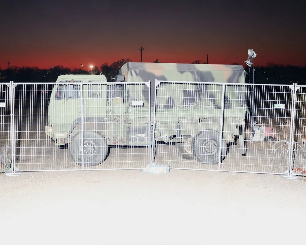 It's Not the Wind by Jordan Conway, photographed by Jordan Conway — A camouflage National Guard transport waits behind temporary metal fencing under a sky bled deep red, a floodlight rigged at the perimeter.