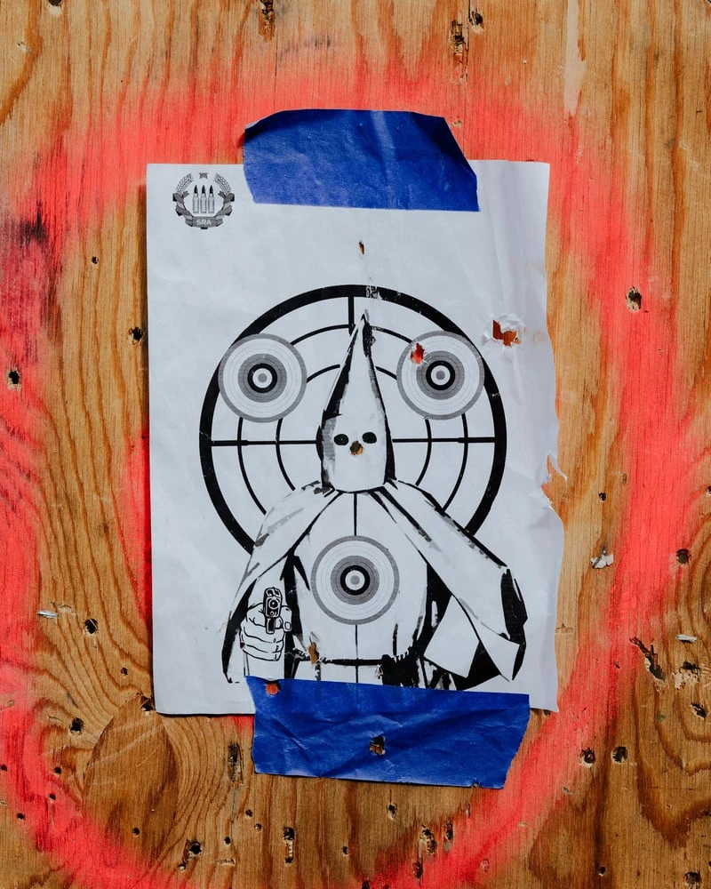 It's Not the Wind by Jordan Conway, photographed by Jordan Conway — A paper shooting target printed with a hooded Klansman raising a pistol, taped to bullet-chewed plywood ringed with red spray paint.