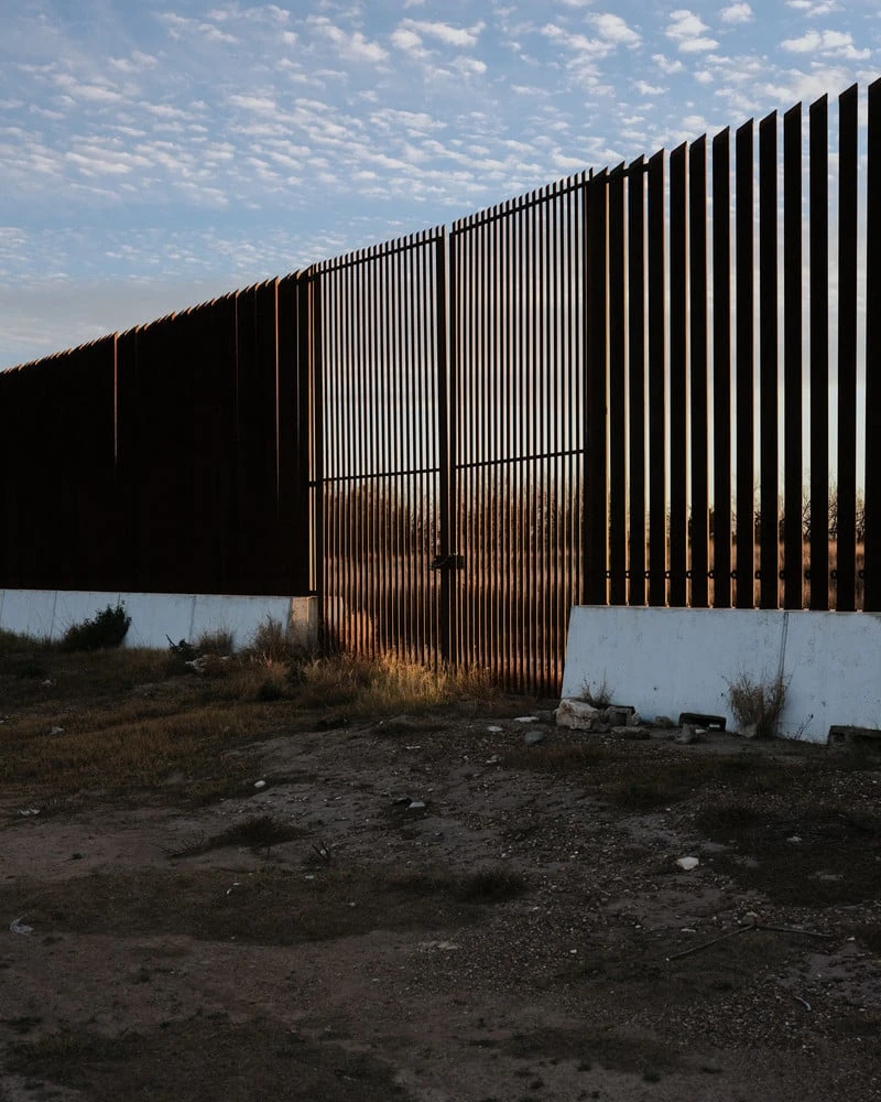 It's Not the Wind by Jordan Conway, photographed by Jordan Conway — A steel bollard border wall runs toward the horizon at dusk, its bars catching gold light above churned, littered ground.