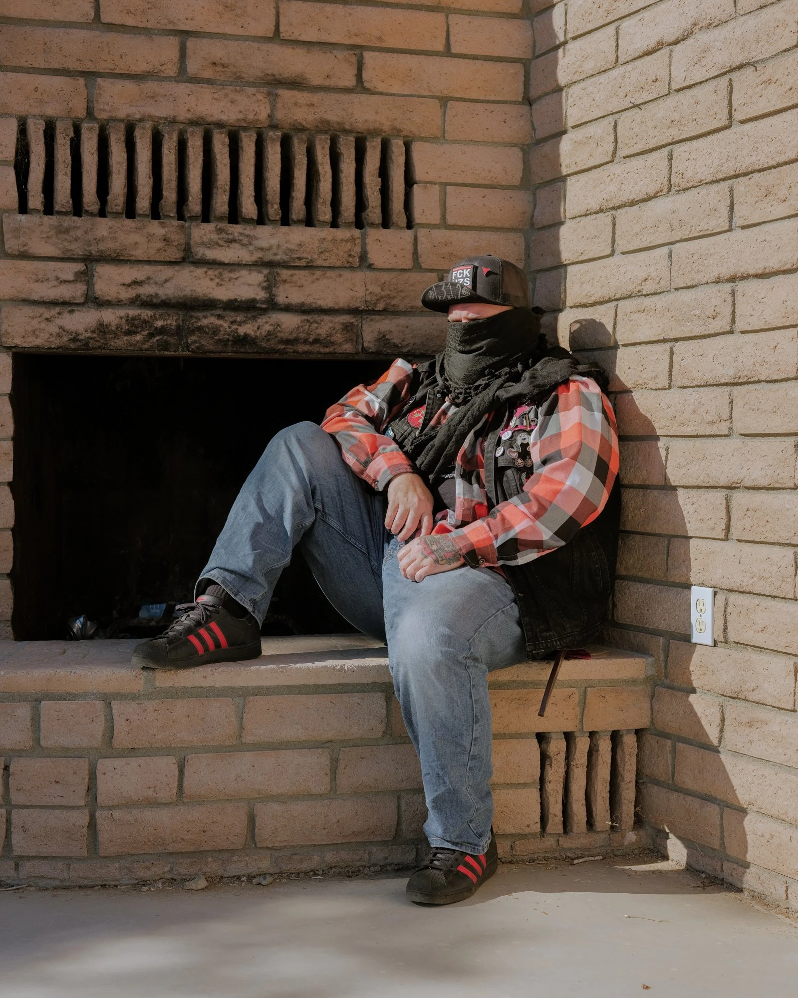 It's Not the Wind by Jordan Conway, photographed by Jordan Conway — A masked man in a plaid flannel shirt and tactical vest sits inside a brick fireplace nook, cap low, boots marked with red stripes.