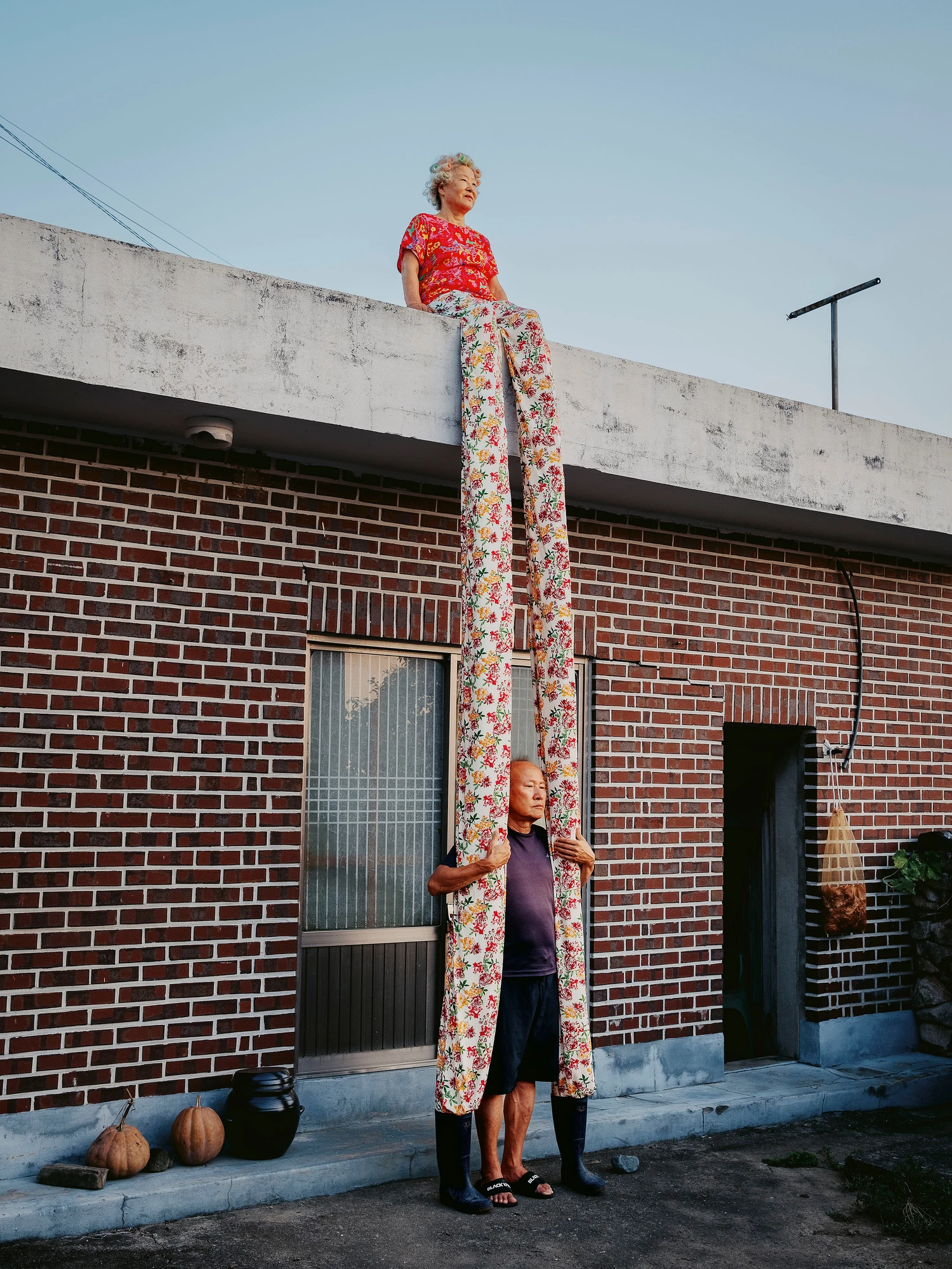 Nirvana by Daesung Lee, photographed by Daesung Lee — Floral trousers run impossibly long from a woman seated on a brick rooftop down into the hands of a man waiting on the ground below.