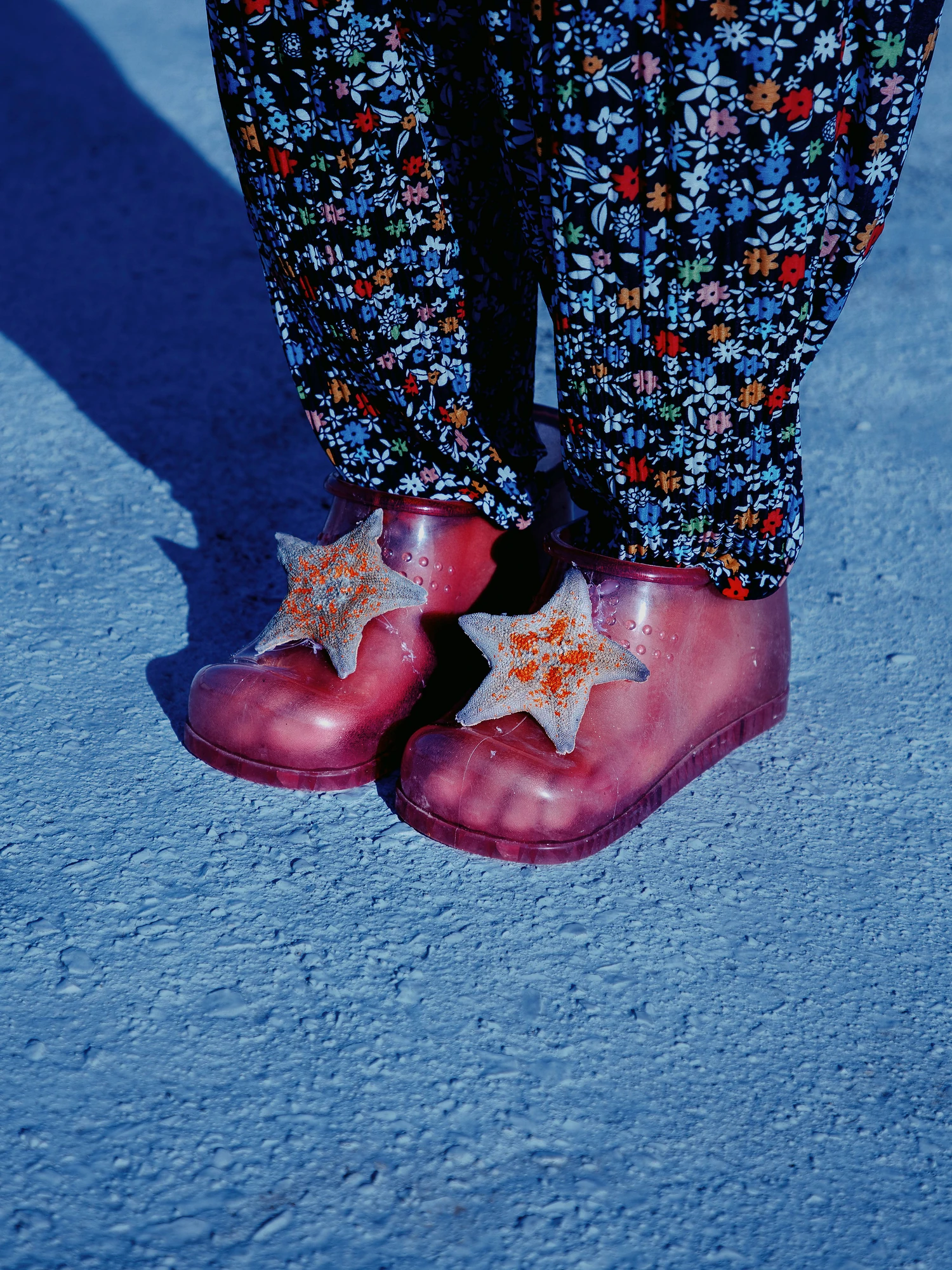 Nirvana by Daesung Lee, photographed by Daesung Lee — Feet in floral trousers wear translucent red garden clogs topped with starfish, casting blue shadows on pale concrete.