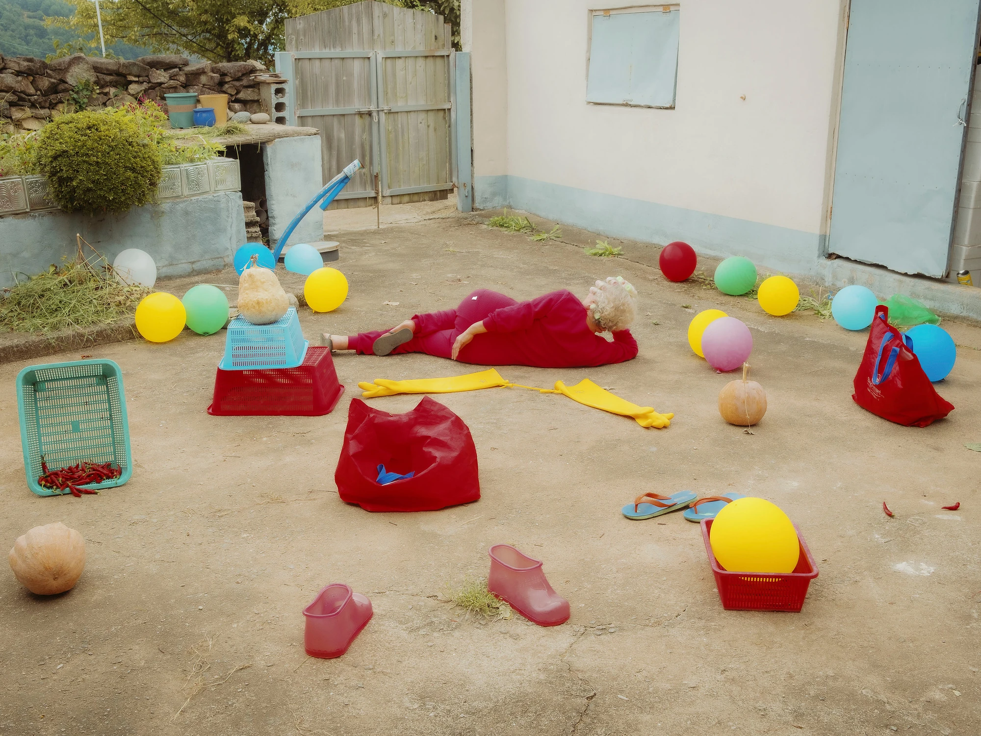 Nirvana by Daesung Lee, photographed by Daesung Lee — A figure in red lies flat in a concrete courtyard scattered with balloons, baskets, garden clogs and pumpkins.