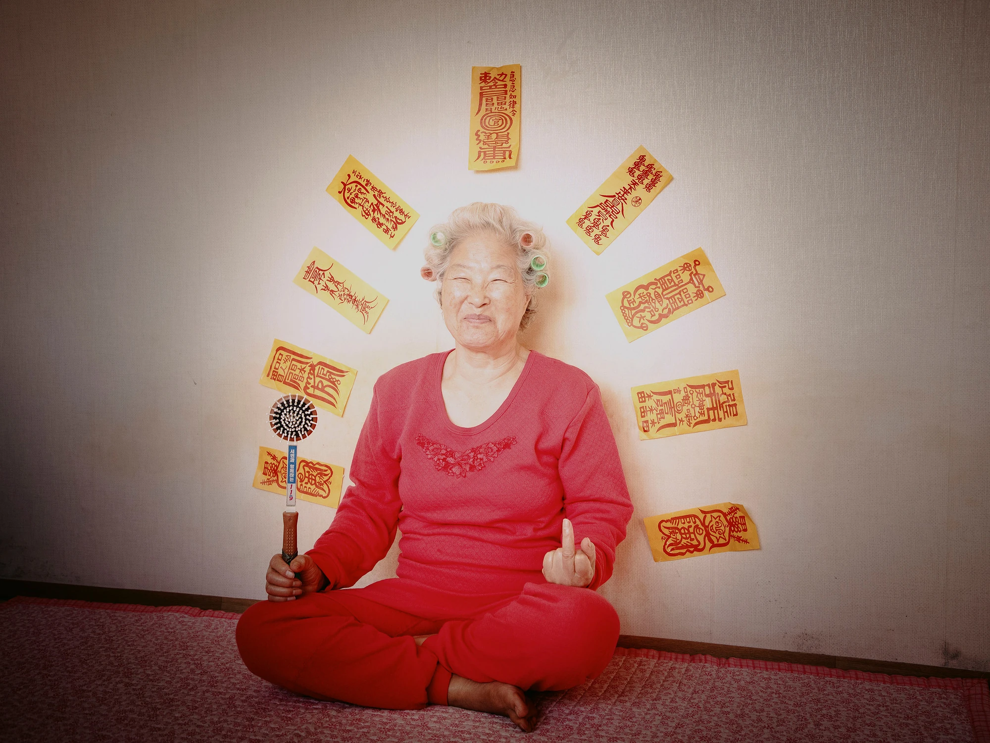 Nirvana by Daesung Lee, photographed by Daesung Lee — A woman sits cross-legged in red, ringed by an arc of yellow shamanic talismans arranged like rays of a rising sun.